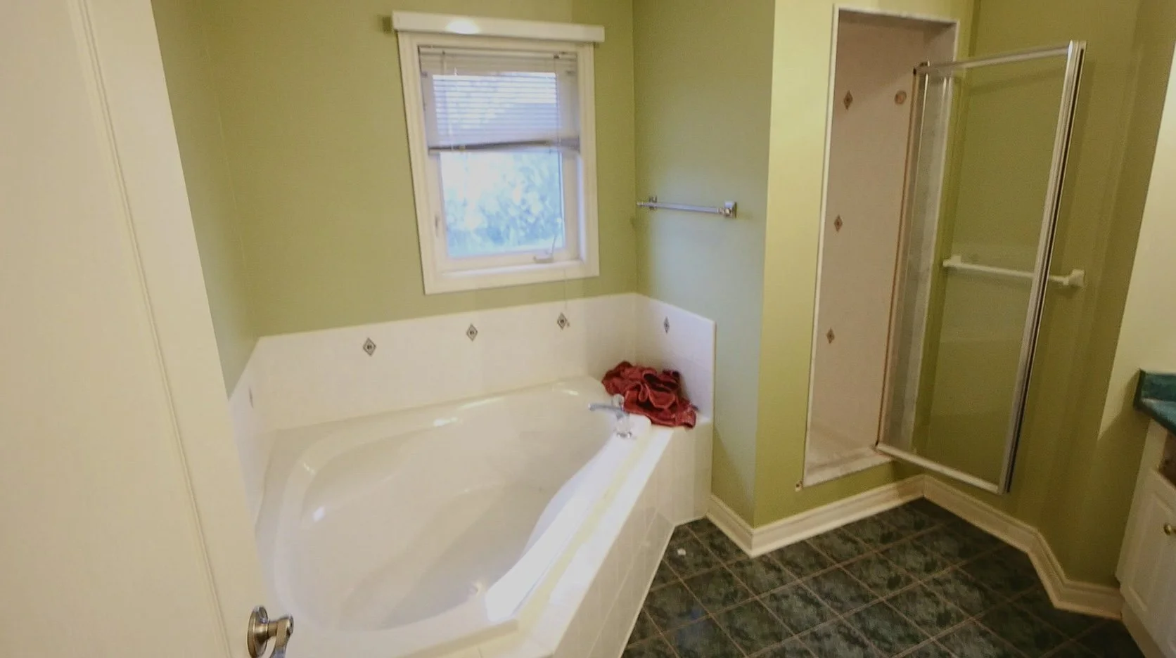 A bathroom with a green wall, a window with blinds, a bathtub with a red towel on the edge, a glass shower stall, a small corner cabinet, and dark tiled flooring.