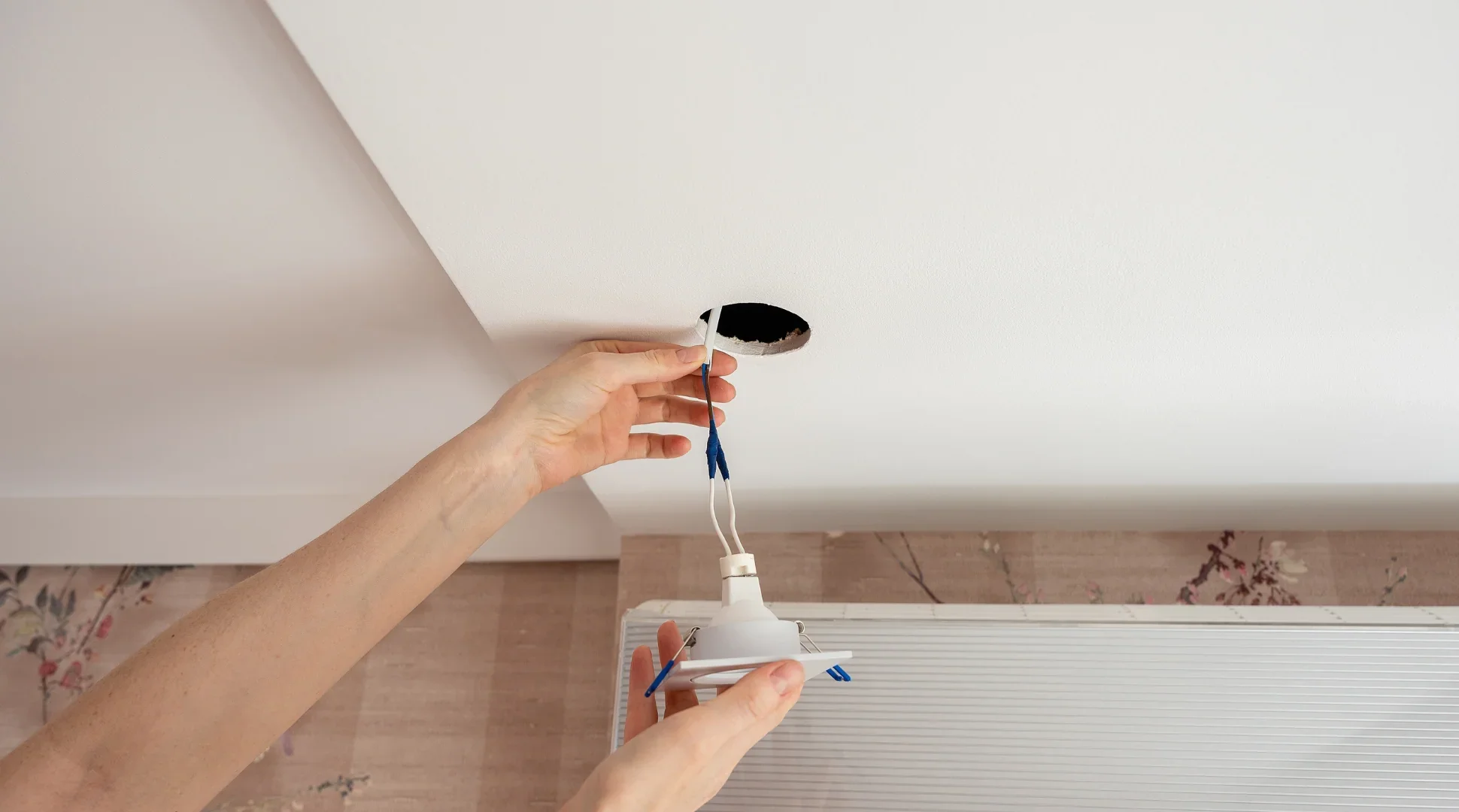 A person installing a ceiling light fixture, holding electrical wires and the fixture near a hole in the ceiling.