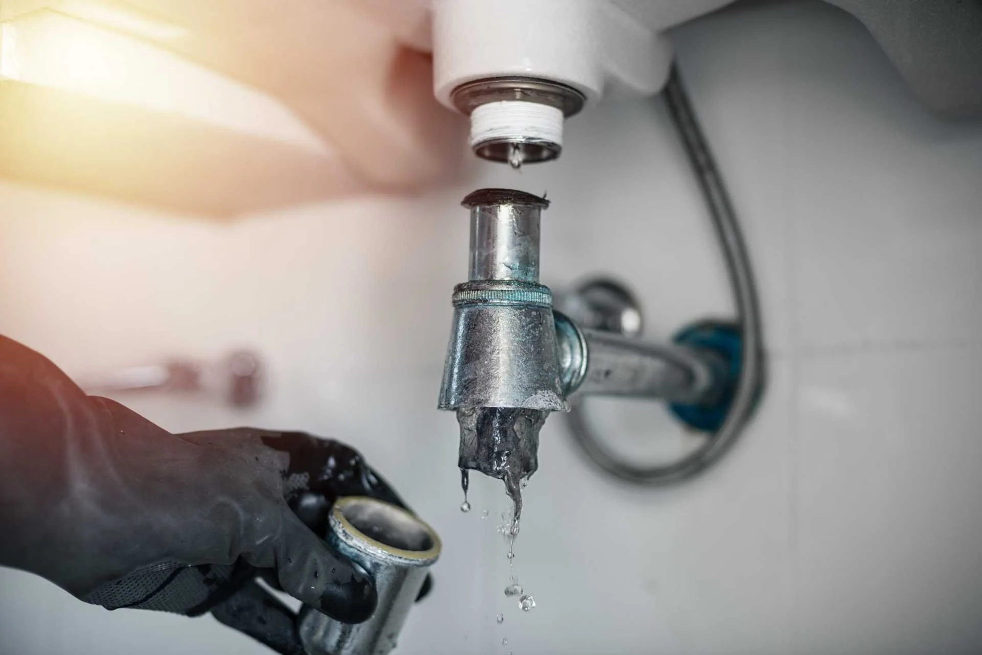 A close-up of a sink faucet with water dripping from it, and a person's hand wearing a black glove holding a pipe fitting underneath the faucet.