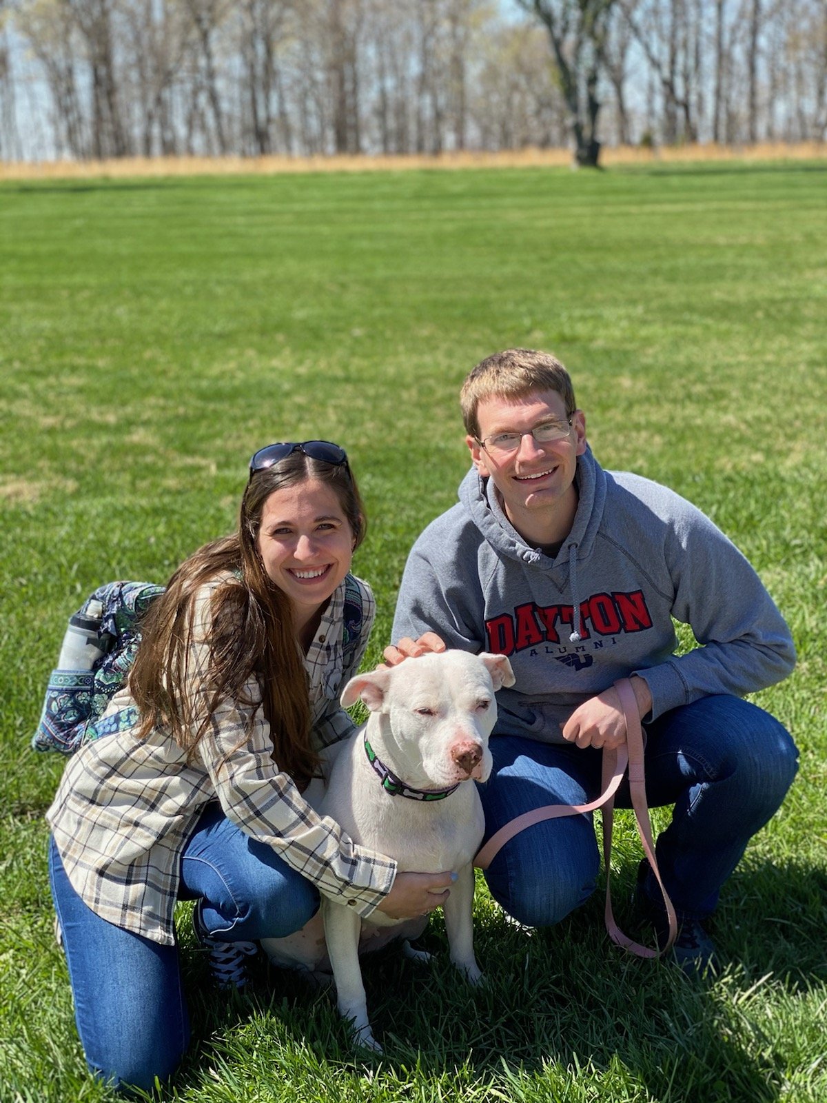 Two smiling people, a woman and a man, crouch on a grassy field with a white dog between them. The woman has long brown hair, sunglasses on her head, and is wearing a plaid shirt and blue jeans. The man has short hair, glasses, and is wearing a gray