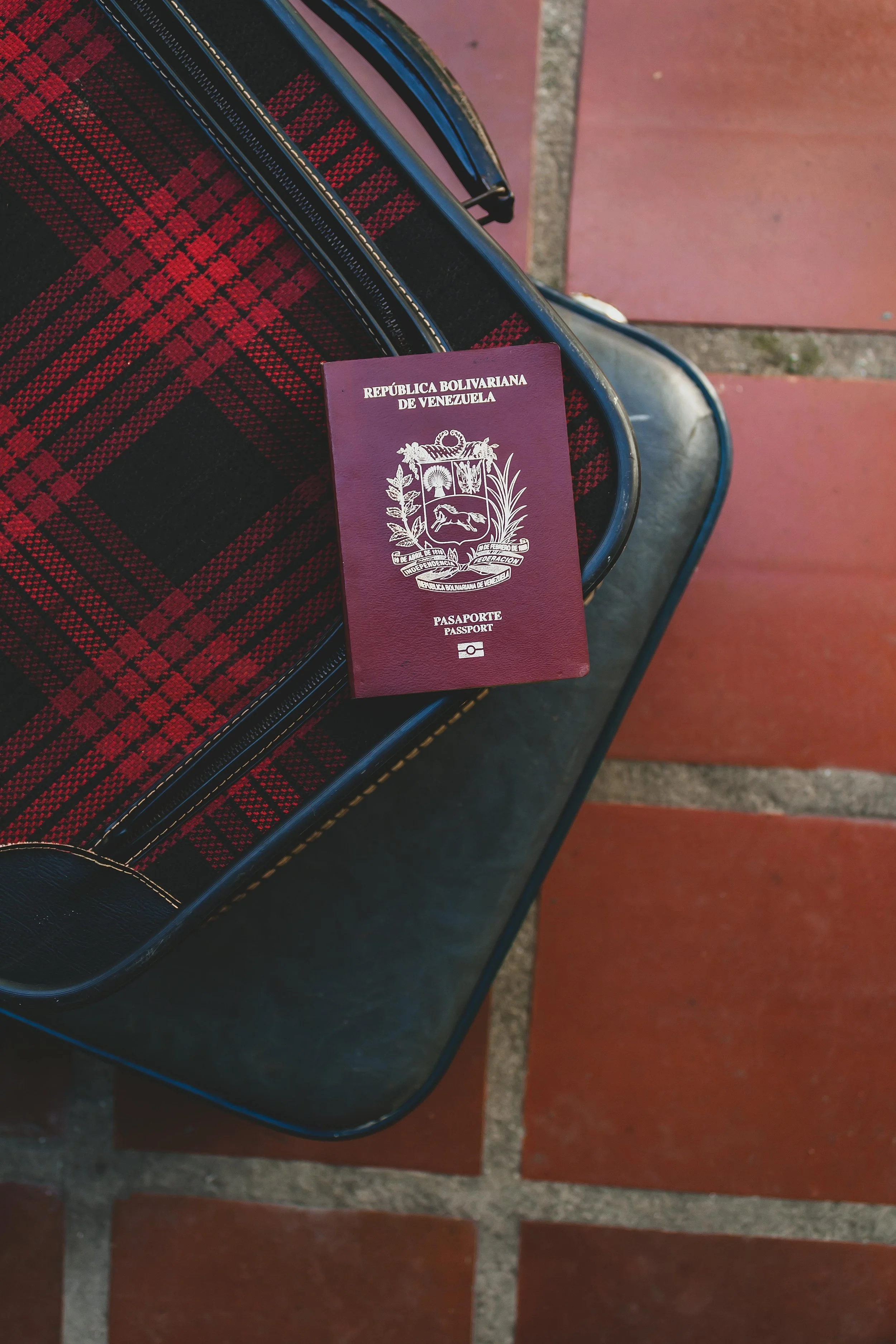 Venezuelan passport resting on a black and red plaid suitcase on a tiled floor.