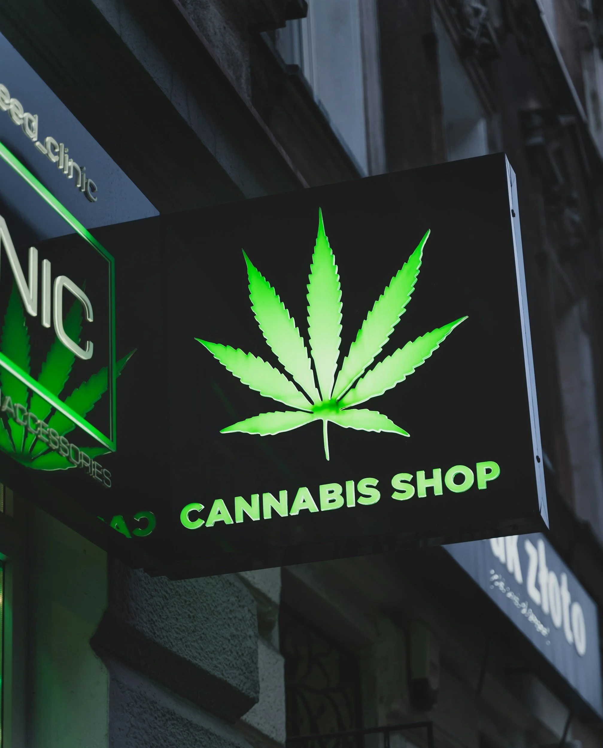 Neon sign of a cannabis leaf with the words "Cannabis Shop" underneath, illuminated in green, on a dark exterior wall.