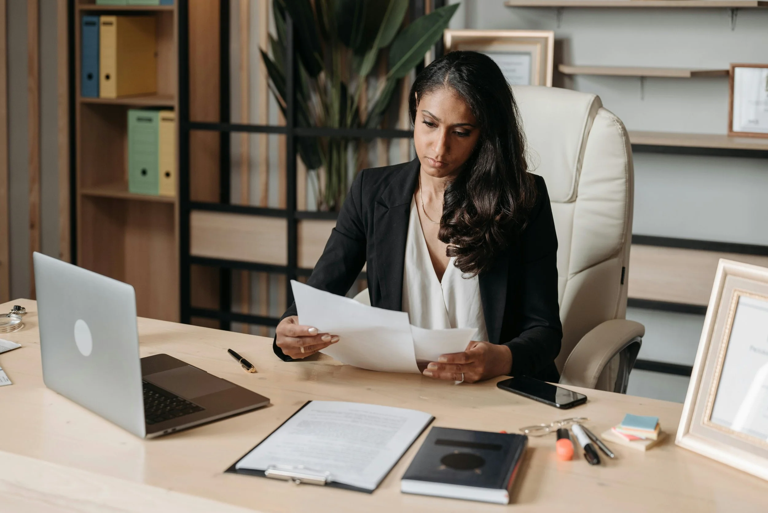 A woman in a black blazer sits at an office desk reviewing papers, with a laptop, smartphone, and office supplies around her.