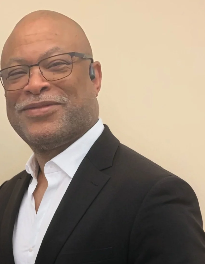 A middle-aged man wearing glasses, a white dress shirt, and a black suit jacket, smiling in front of a plain light-colored wall.