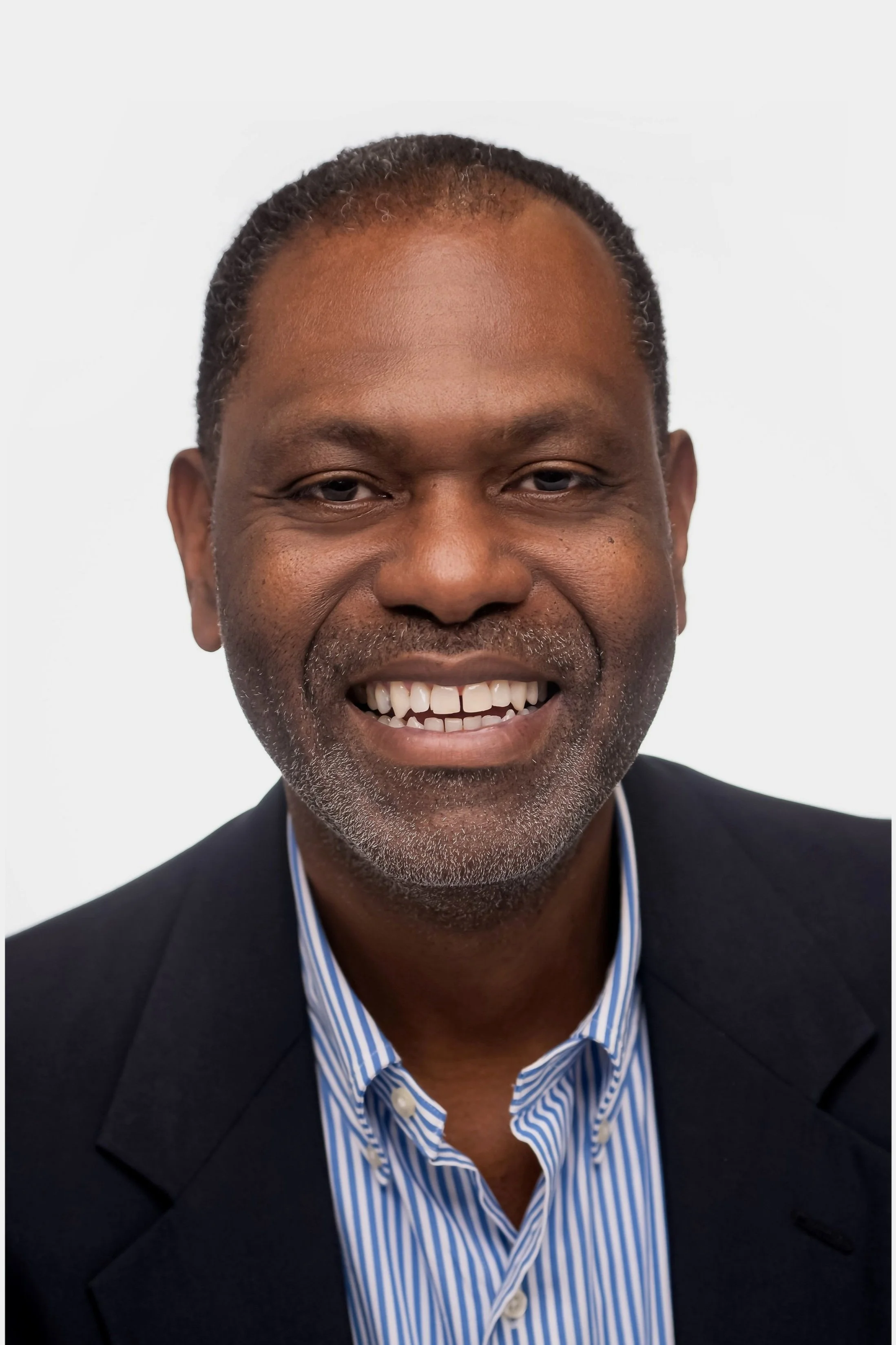 Close-up portrait of a smiling African American man with short hair, a beard, wearing a dark suit jacket and a blue and white striped shirt against a white background.