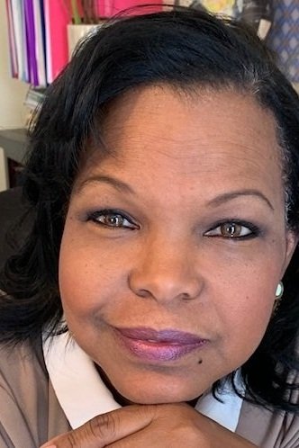 Close-up of a woman with dark hair, wearing makeup, smiling softly at the camera, with a background of office shelves and binders.