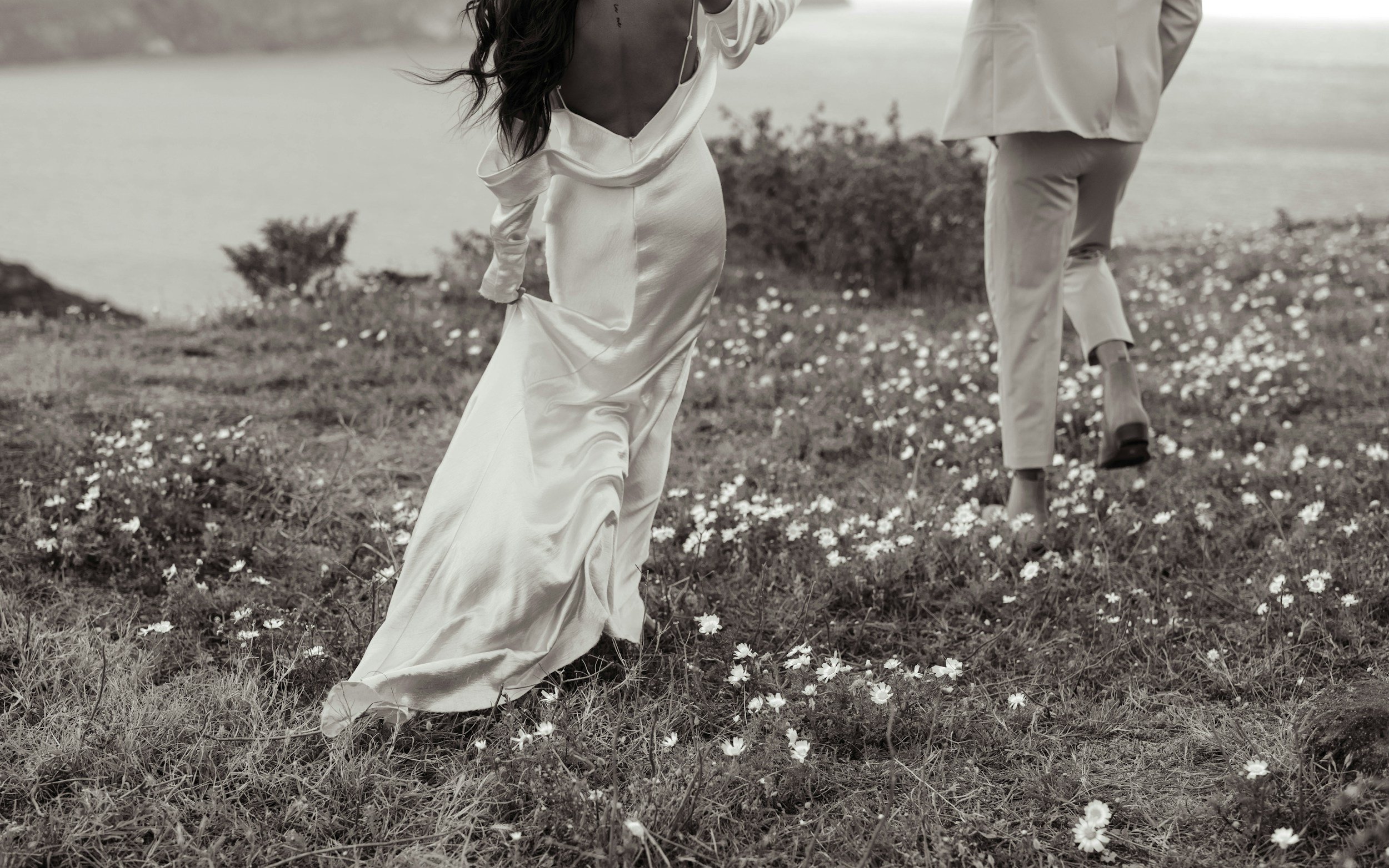 A woman in a long, flowing satin dress with an open back and a man in a light-colored suit walking through a field of small daisies near a body of water.