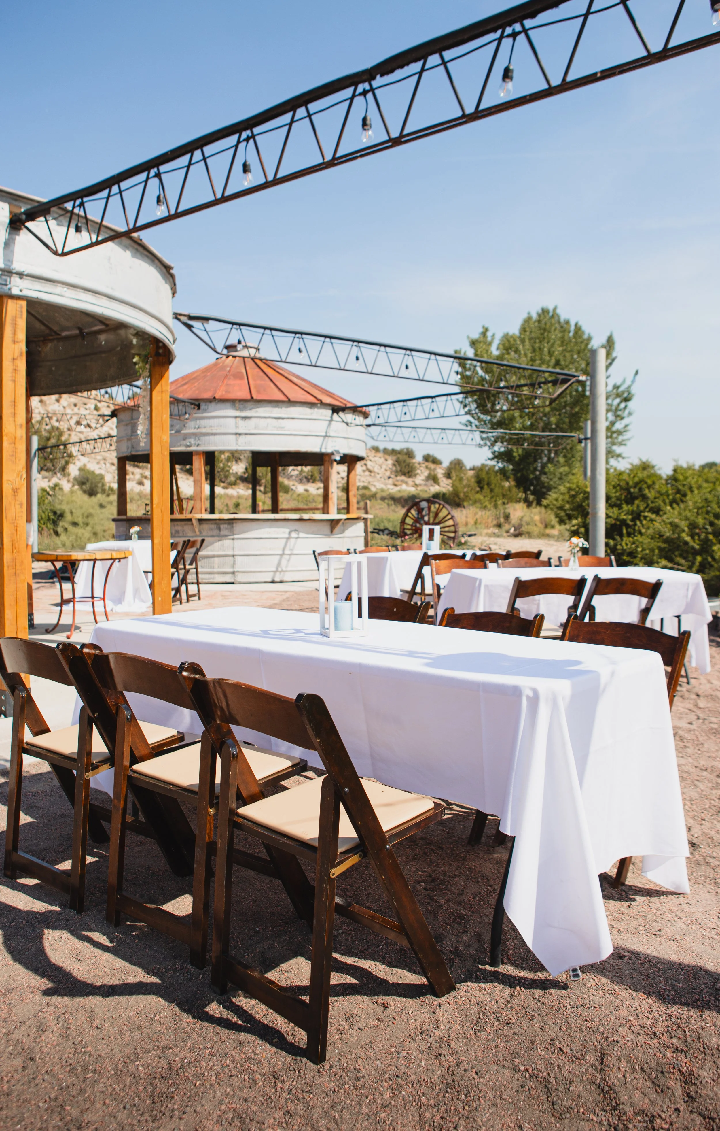 Outdoor dining setup with tables covered in white tablecloths and wooden chairs on a gravel surface, in front of wooden and metal structures with scenic landscape and trees in the background.