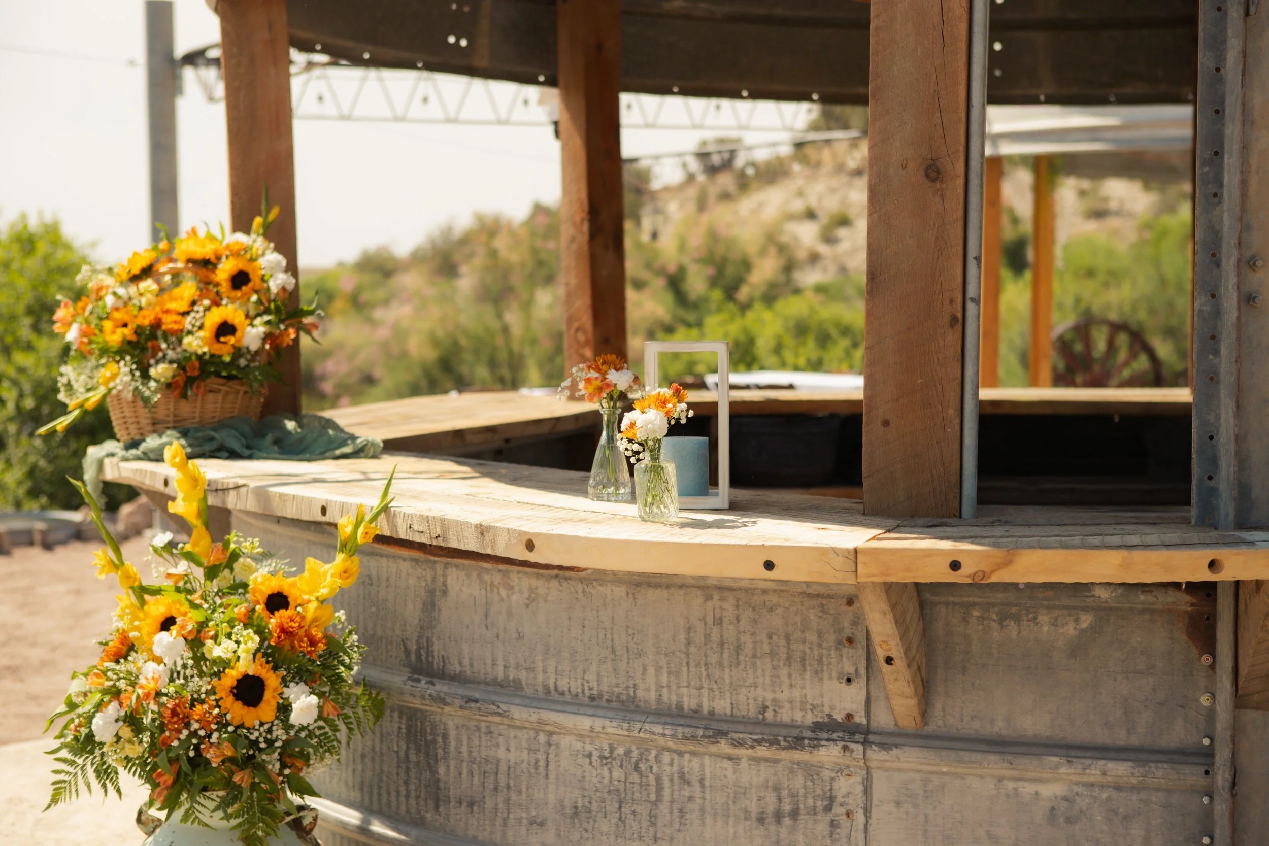 A wooden and metal structure with floral arrangements including sunflowers and other flowers placed on a ledge and in a basket, set outdoors during daytime.
