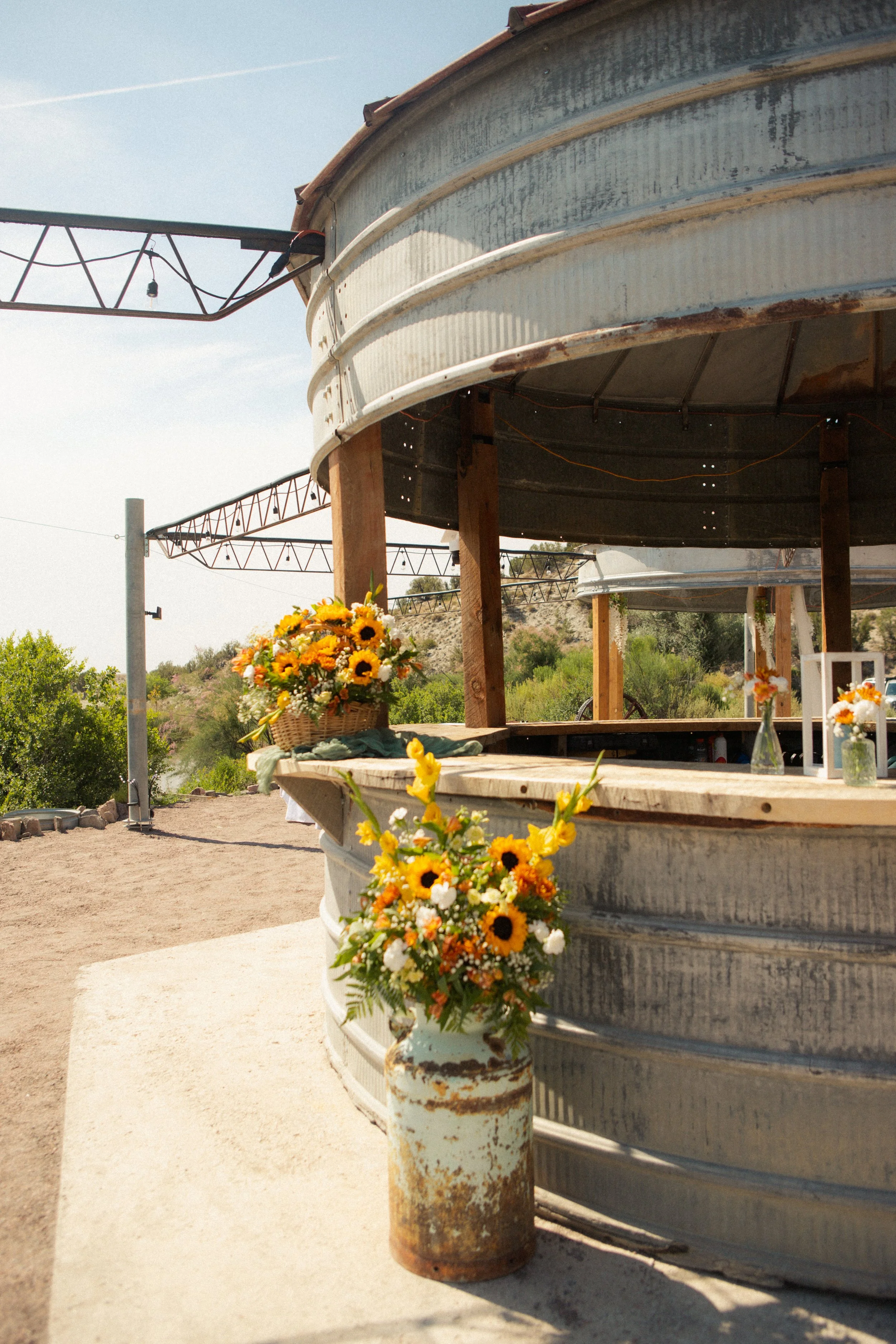 Decorative flowers in a basket and a rusty metal container are placed near a large, corrugated, weathered metal structure. The setting appears outdoor with a clear sky and hills in the background.