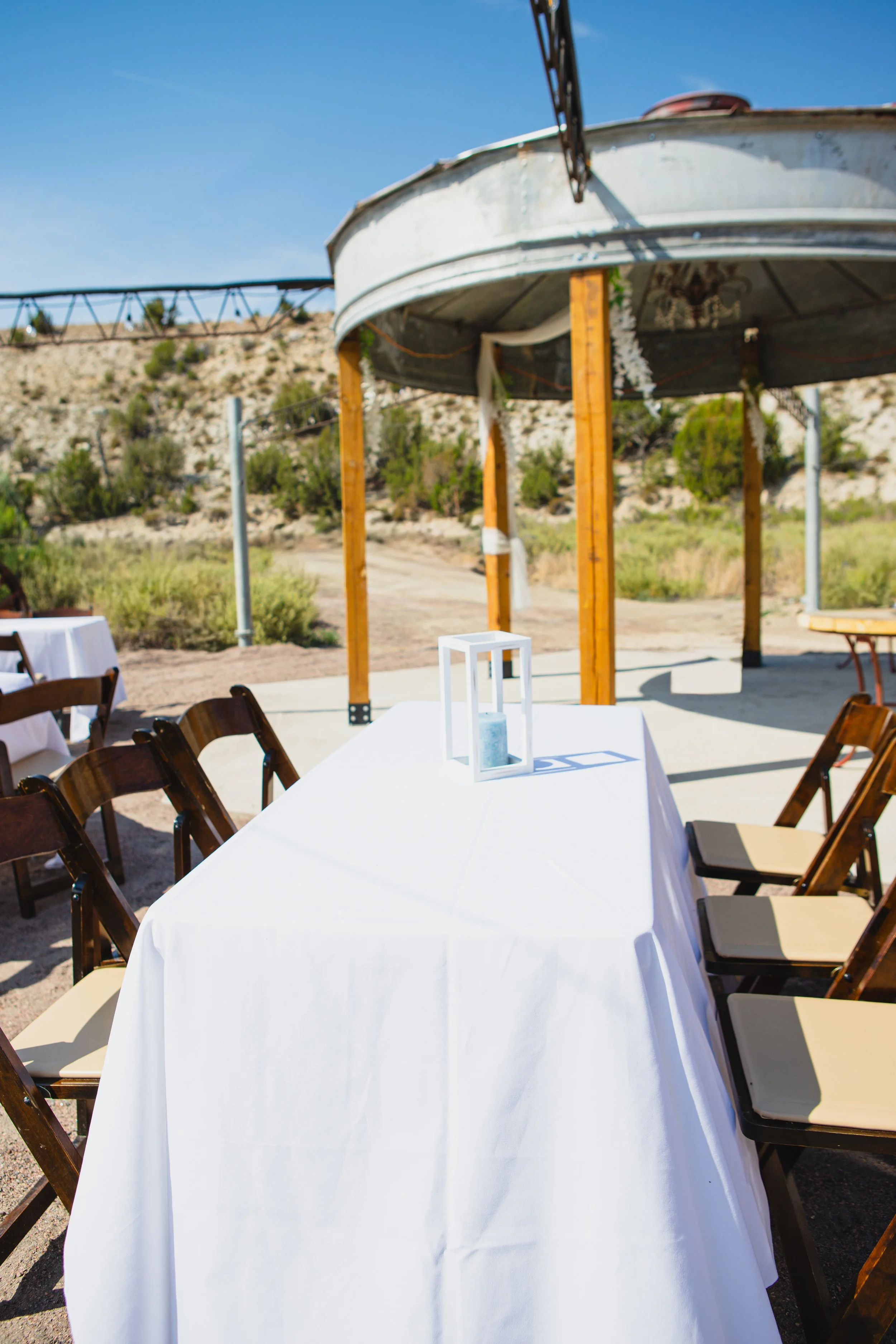 Outdoor dining setup with a long white tablecloth-covered table, wooden chairs, and a tent structure with supporting wooden posts, against desert-like hill and clear blue sky background.