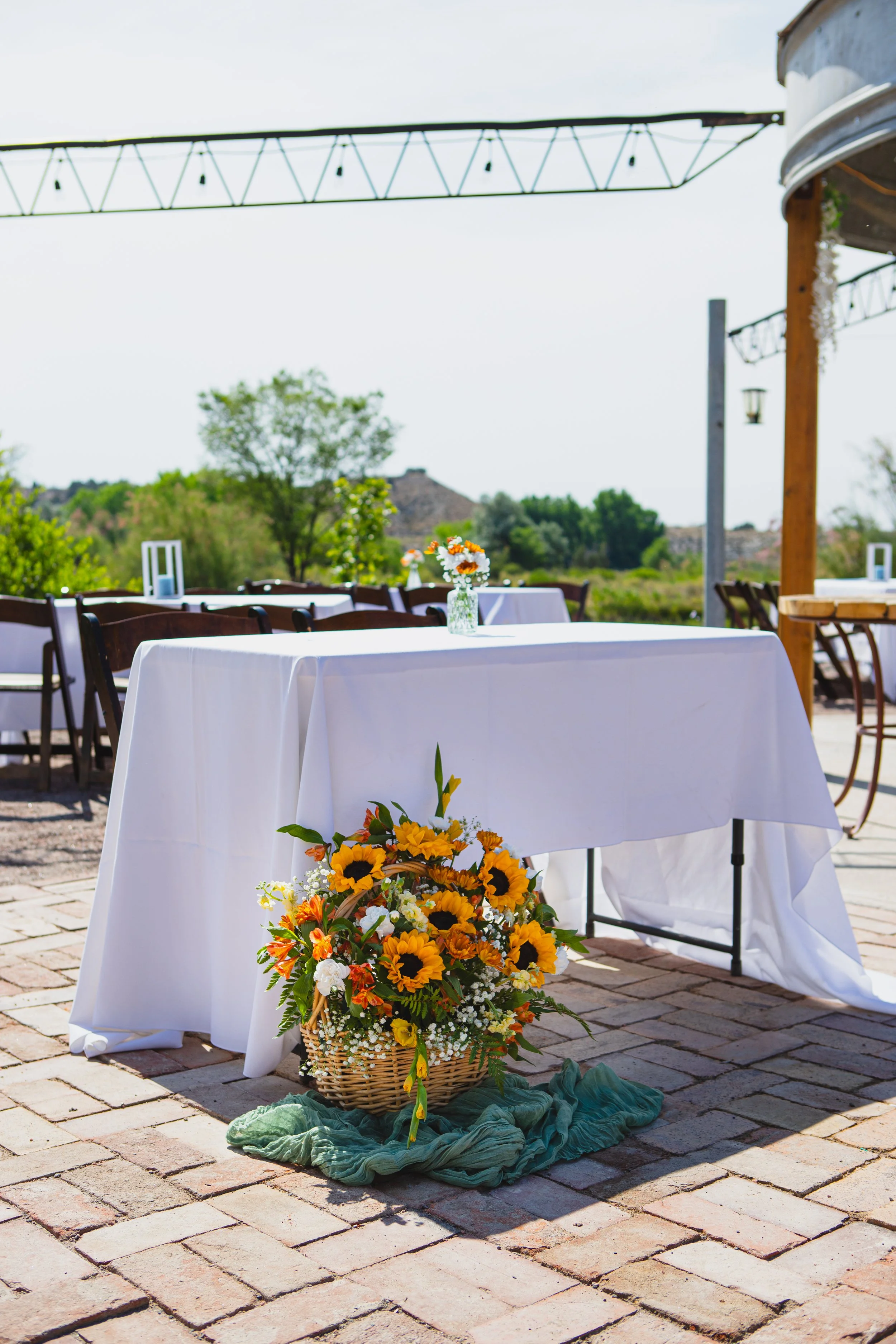 Outdoor outdoor wedding setup with a white tablecloth-covered table, a basket of yellow and white flowers, and a green cloth on cobblestone ground, with chairs and trees in the background.