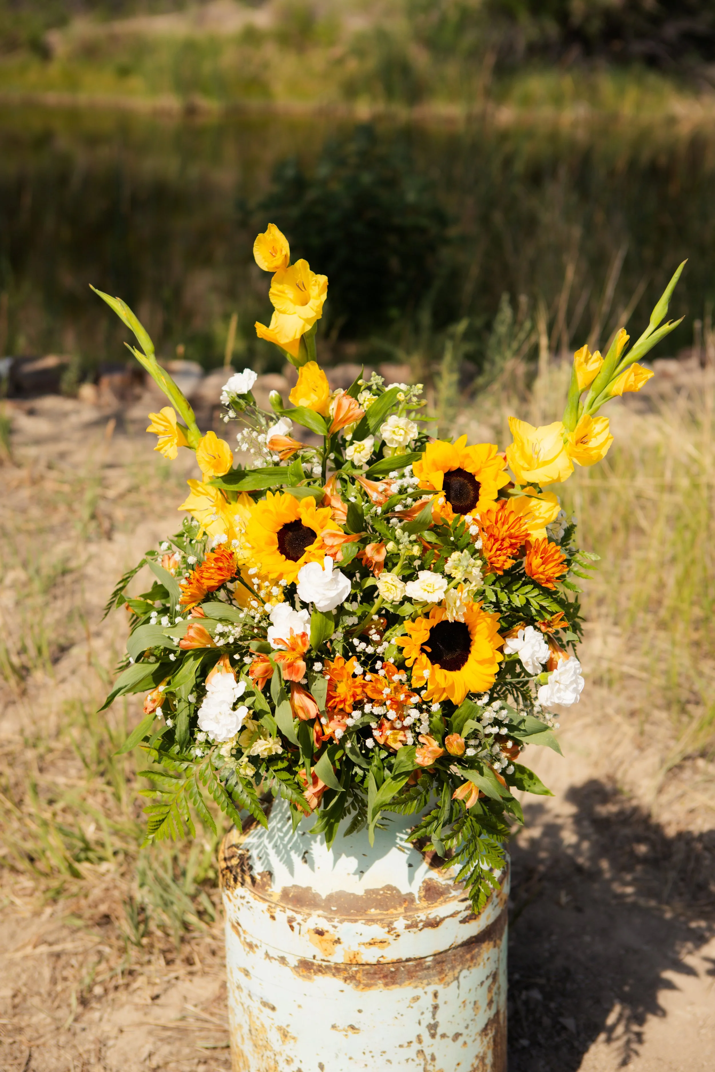 A bouquet of yellow, orange, and white flowers arranged in front of a rusted metal pipe outdoors.
