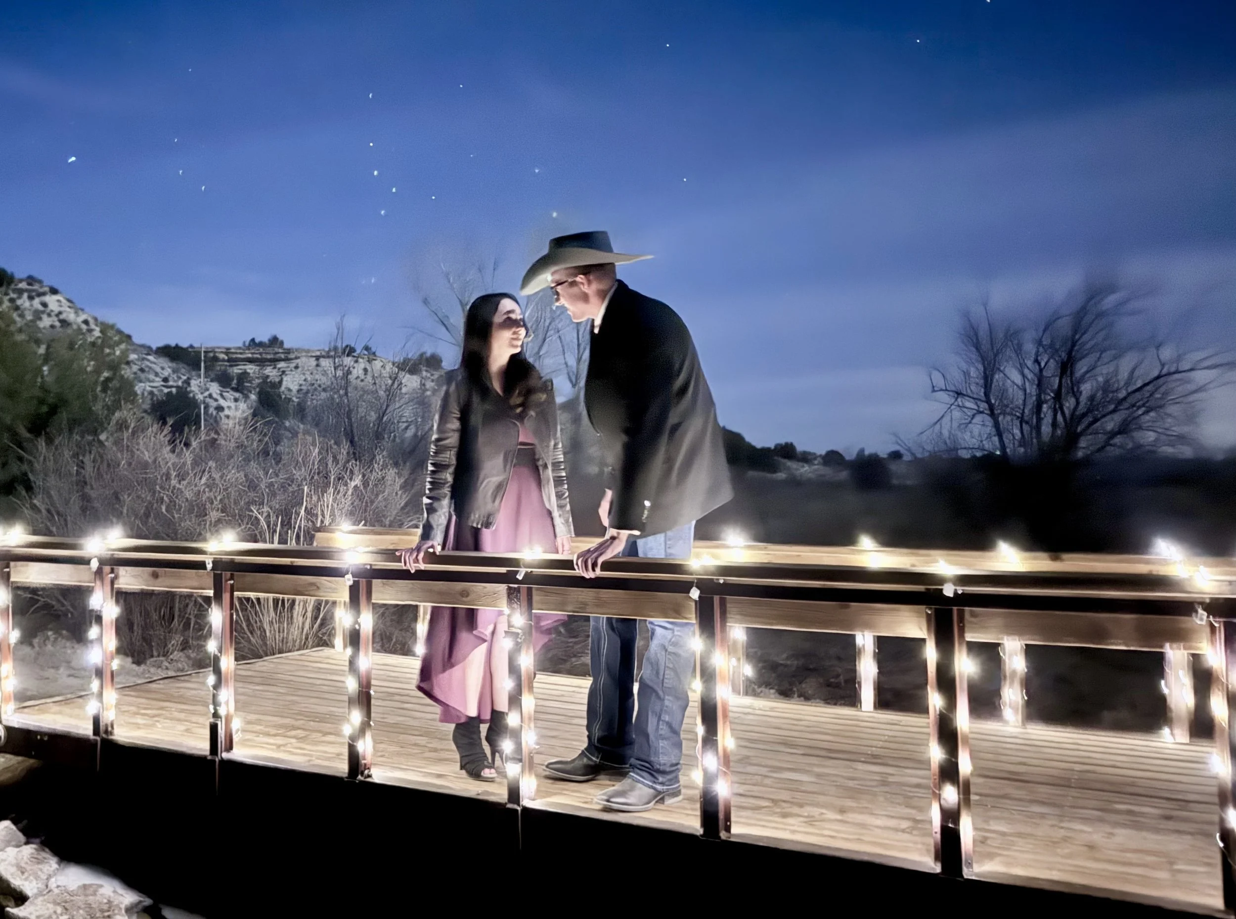 Bride and groom on bridge over stream and waterfall at night. The bridge is covered in twinkle fair lights glowing like fireflies at night, at Lost Pond a rustic outdoor wedding venue near Pueblo Colorado.