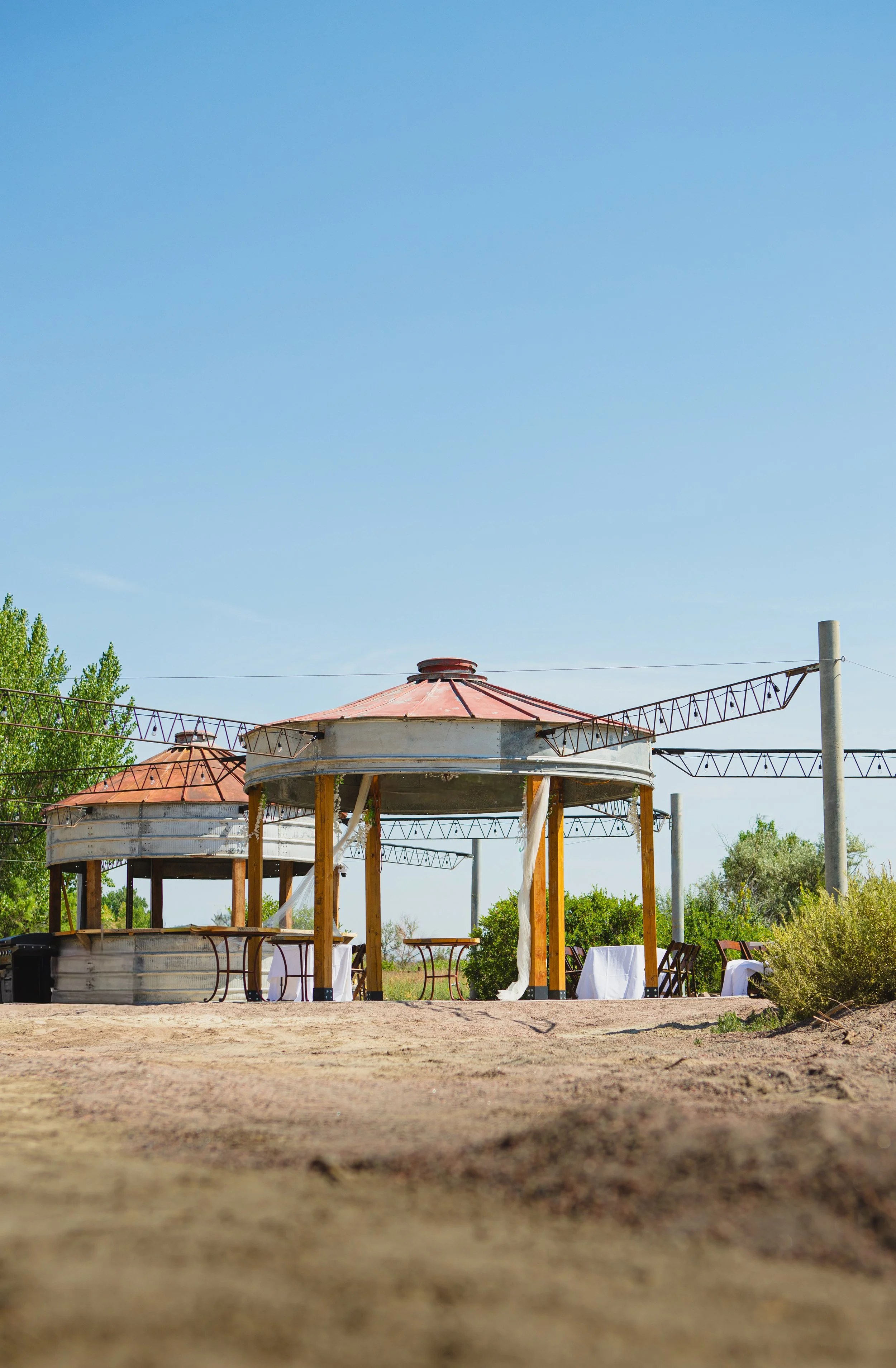 A partially constructed outdoor gazebo with a round metal roof in an open area with a dirt ground and trees in the background, and overhead string lights.