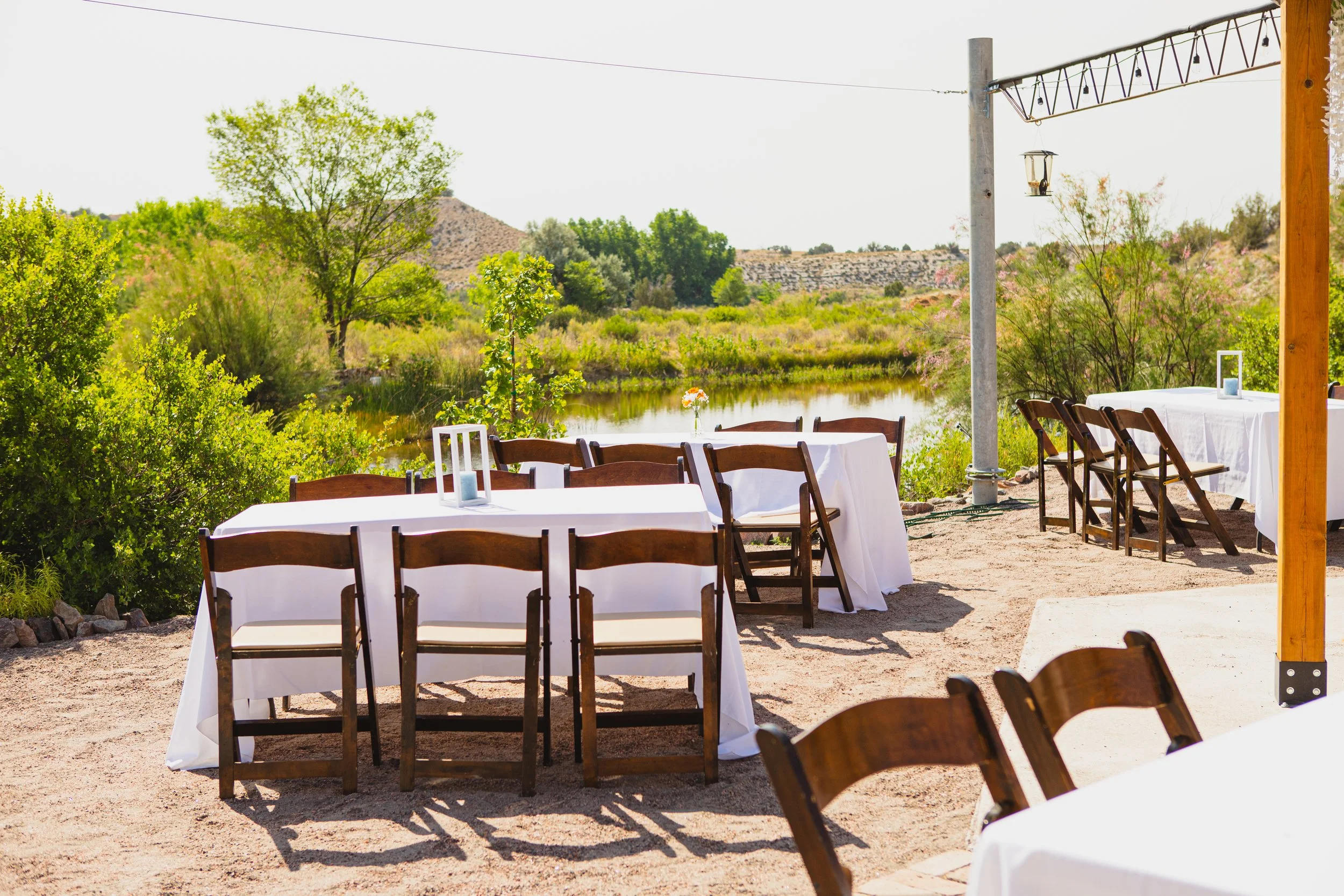 Outdoor dining area with white tablecloth-covered tables and wooden chairs near a river, surrounded by green trees and shrubs, with hills in the distance.