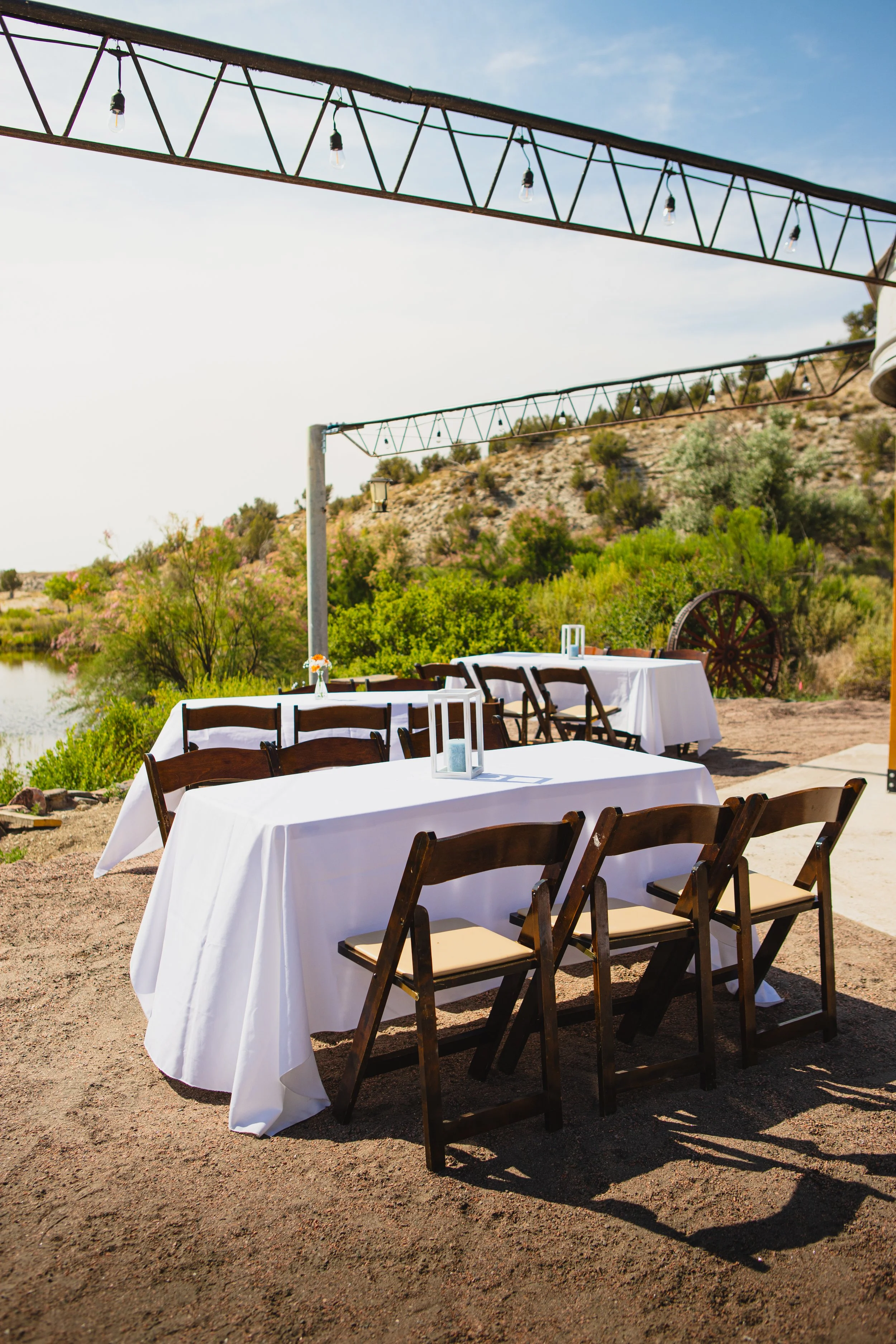 Outdoor reception area with long tables covered in white tablecloths, surrounded by dark wooden chairs with light-colored cushions, set on a dirt ground with green trees and rolling hills in the background, under a blue sky with a few clouds, and str