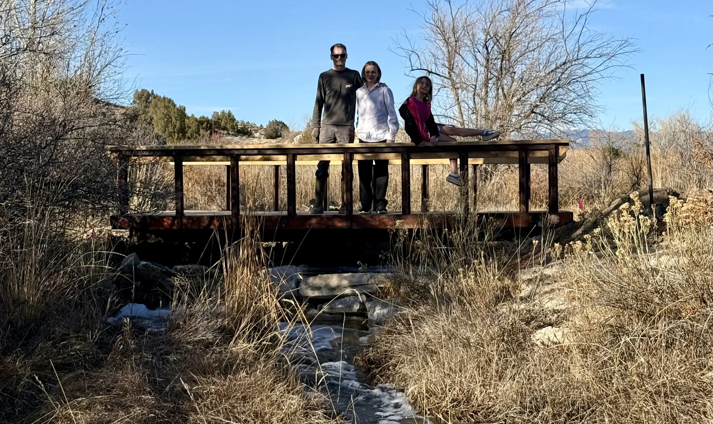 Three people standing on a rustic wooden bridge over a waterfall with mountains in the background. This is at Lost Pond outdoor wedding venue near Pueblo, Colorado.