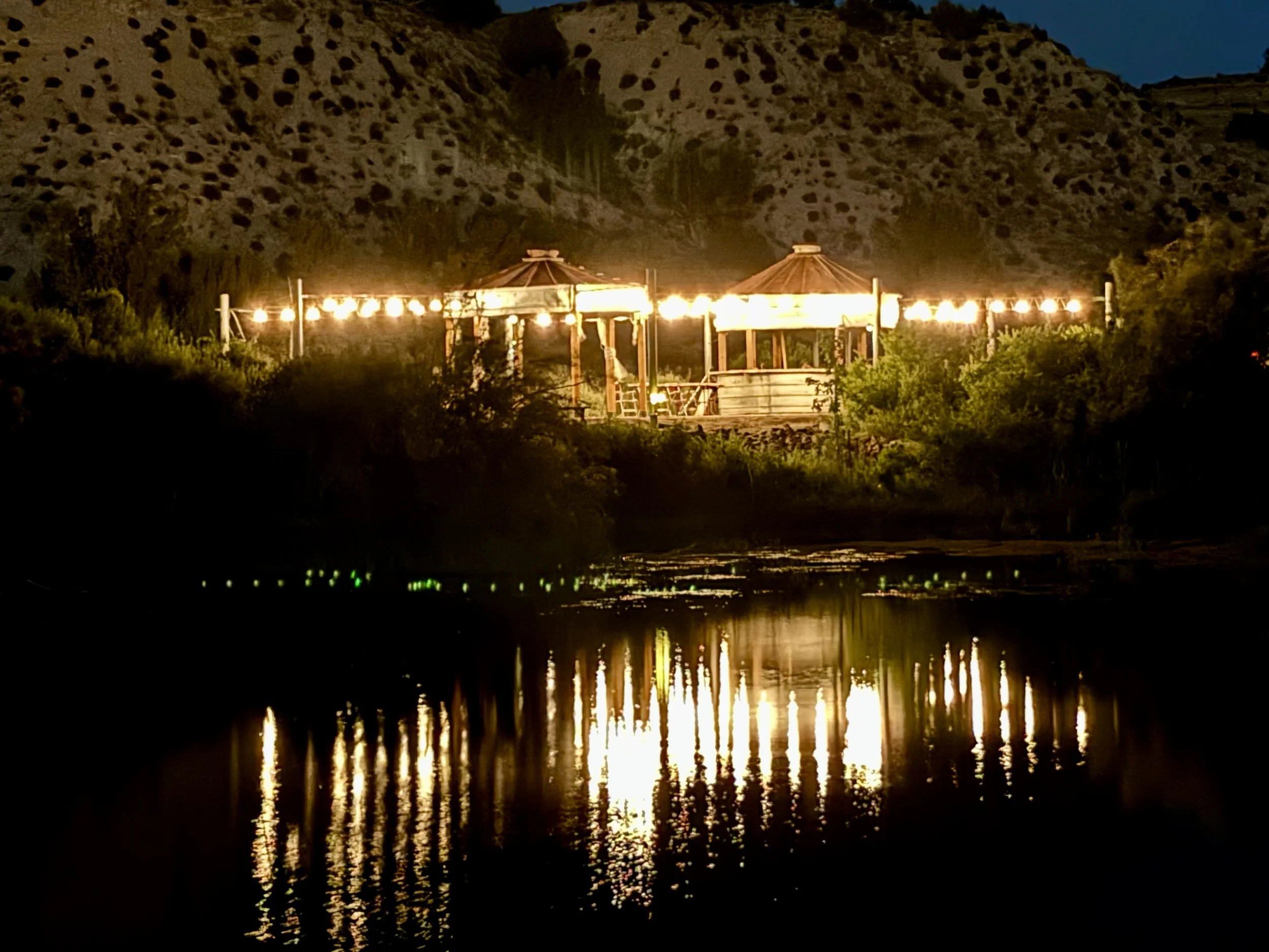 Nighttime scene of a pond with a reflection of string lights, mountains in the background, and a cloudy sky. At Lost Pond outdoor wedding venue near Colorado Springs, Colorado.