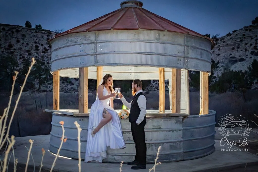 A bride and groom make a champaign toast by a rustic bar silo at their wedding reception at Lost Pond wedding venue near Colorado Springs, Colorado.