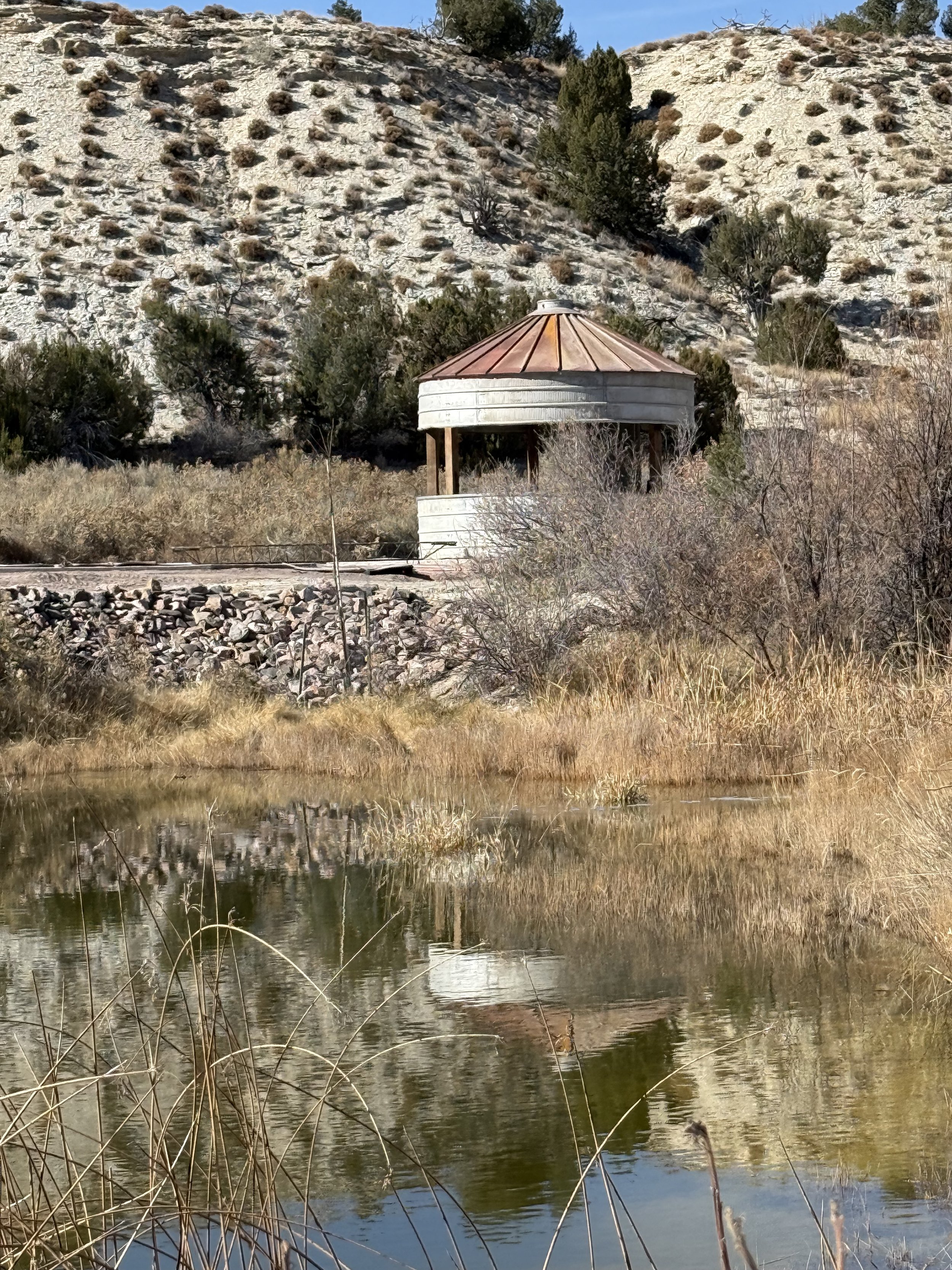 A rustic gazebo with a rust-colored metal roof located near water, with by the Canon City Colorado mountains. Lost Pond wedding venue