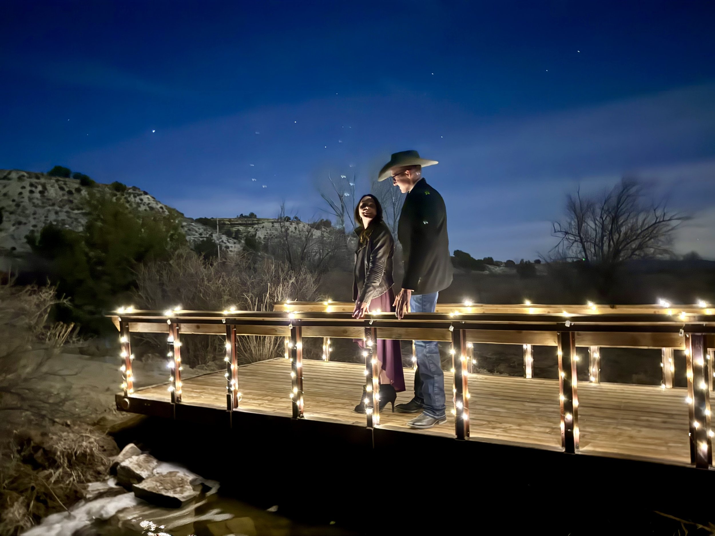 Engaged couple, bride and groom, on bridge lit up with twinkle and string lights under a starlit sky at Lost Pond wedding venue.