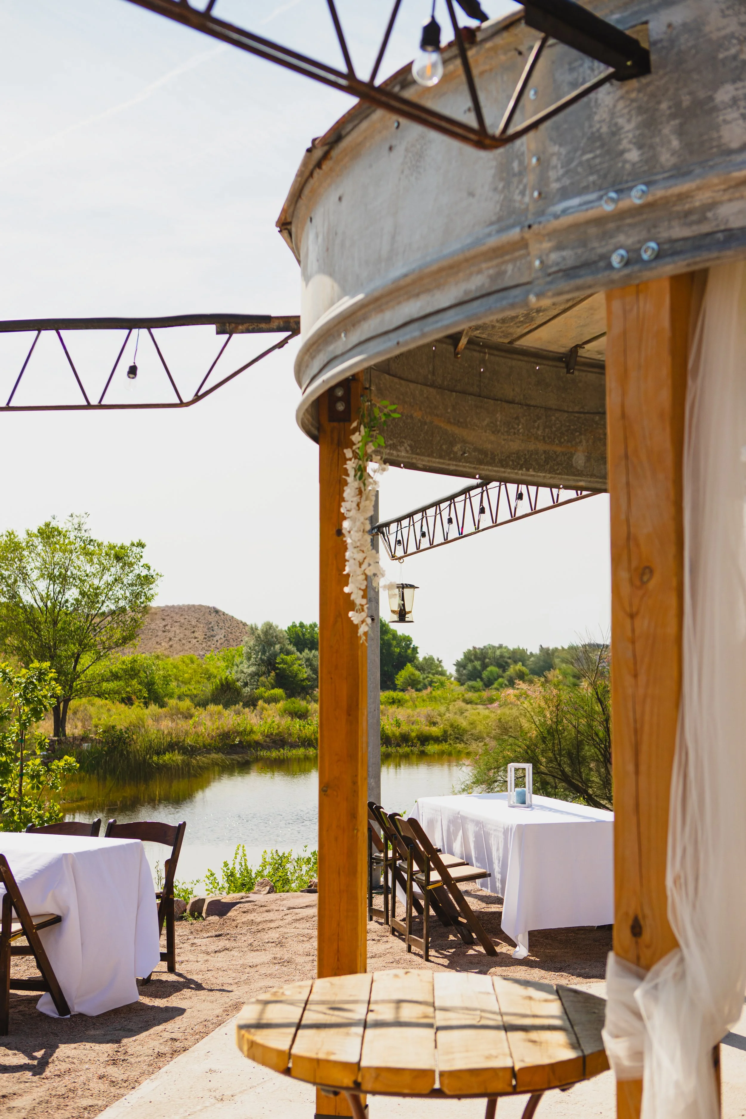 Outdoor restaurant setup with tables covered in white cloth, wooden chairs, and a view of a river, trees, and hills in the background. The scene is shaded by a wooden canopy with decorative string lights.
