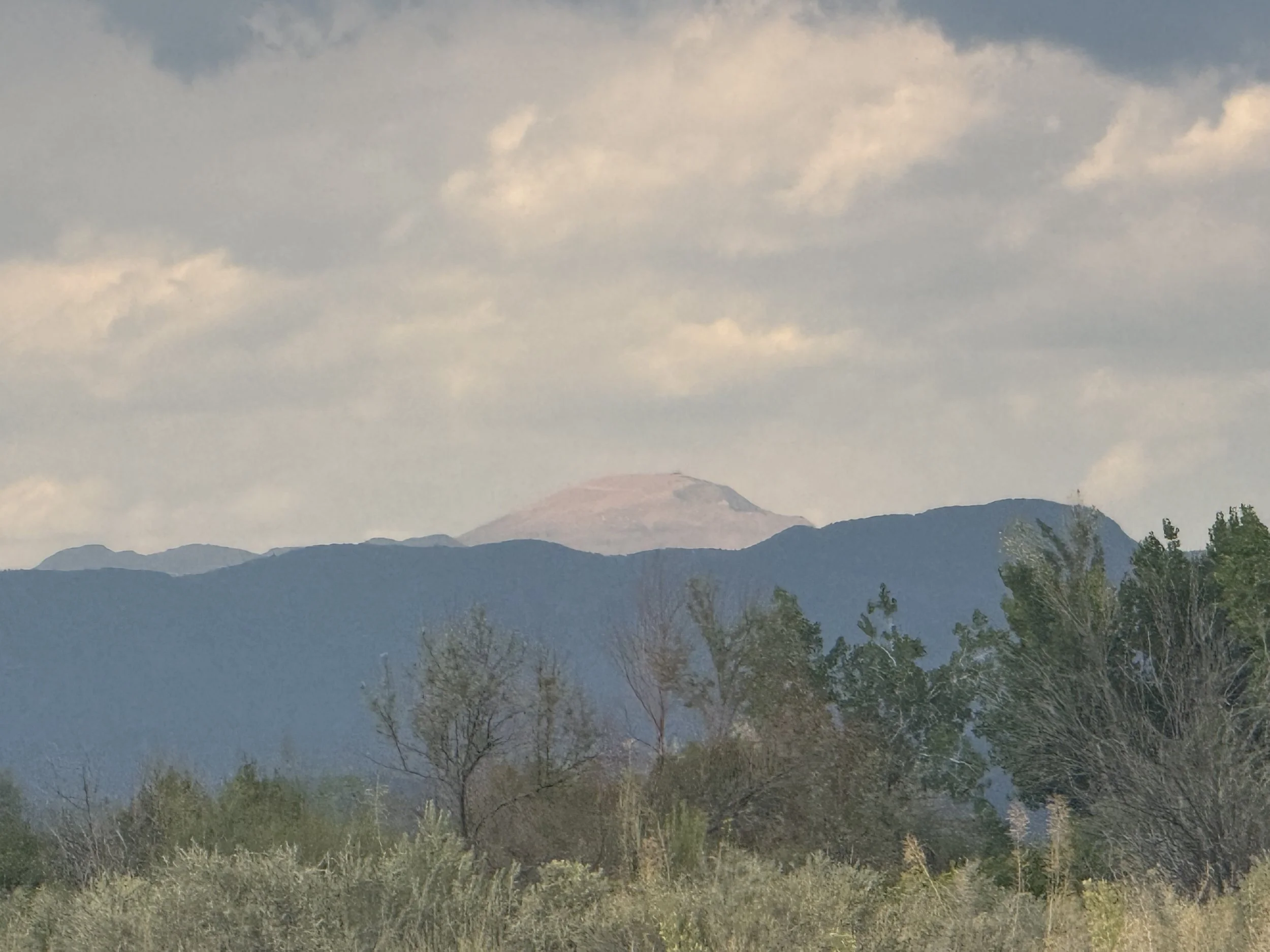 Mountain landscape with cloud-covered sky, distant peaks, and trees in the foreground.