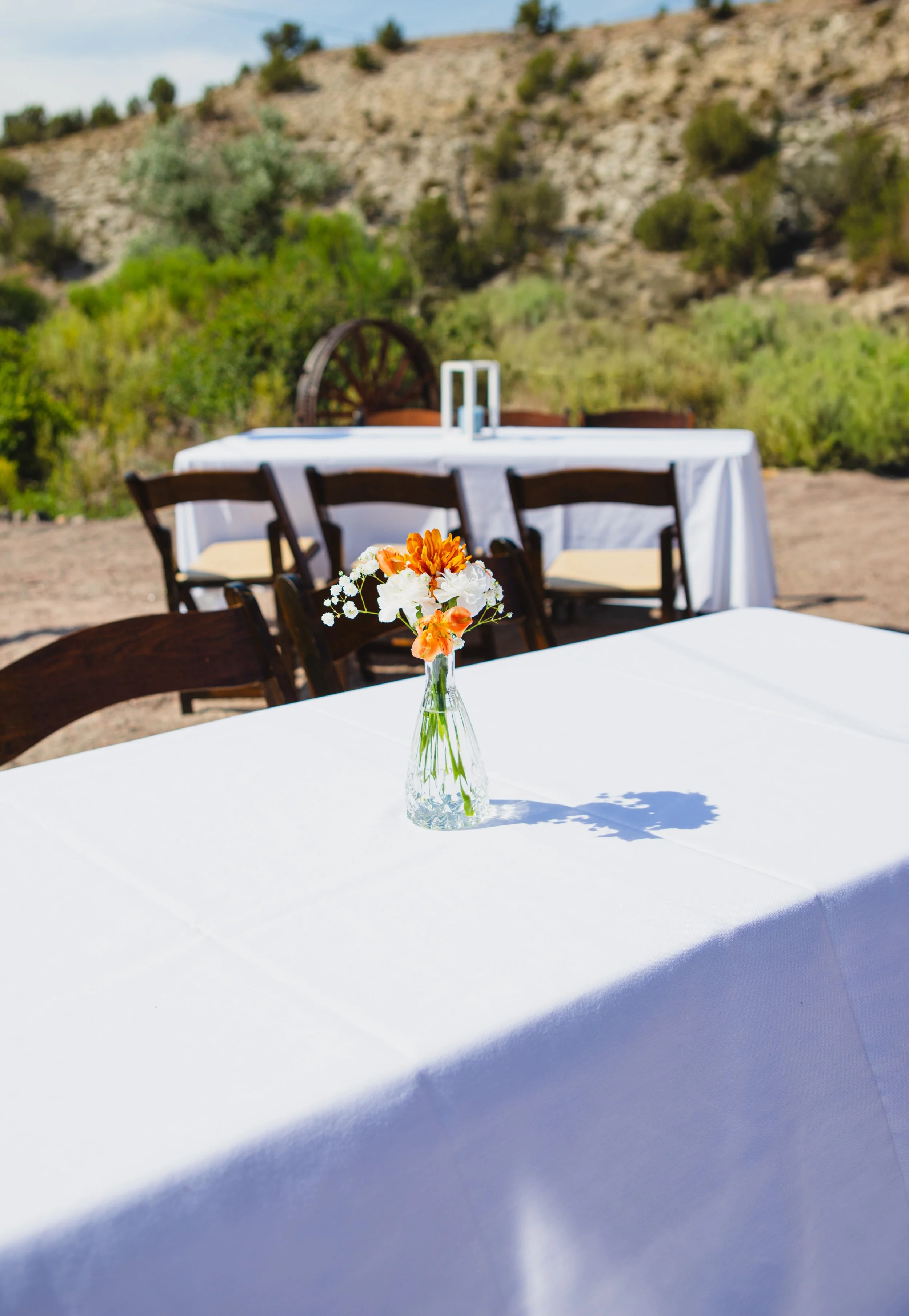 A small glass vase with a bouquet of orange, white, and white flowers on a white tablecloth, with chairs and a scenic outdoor background featuring trees and a rocky hillside.