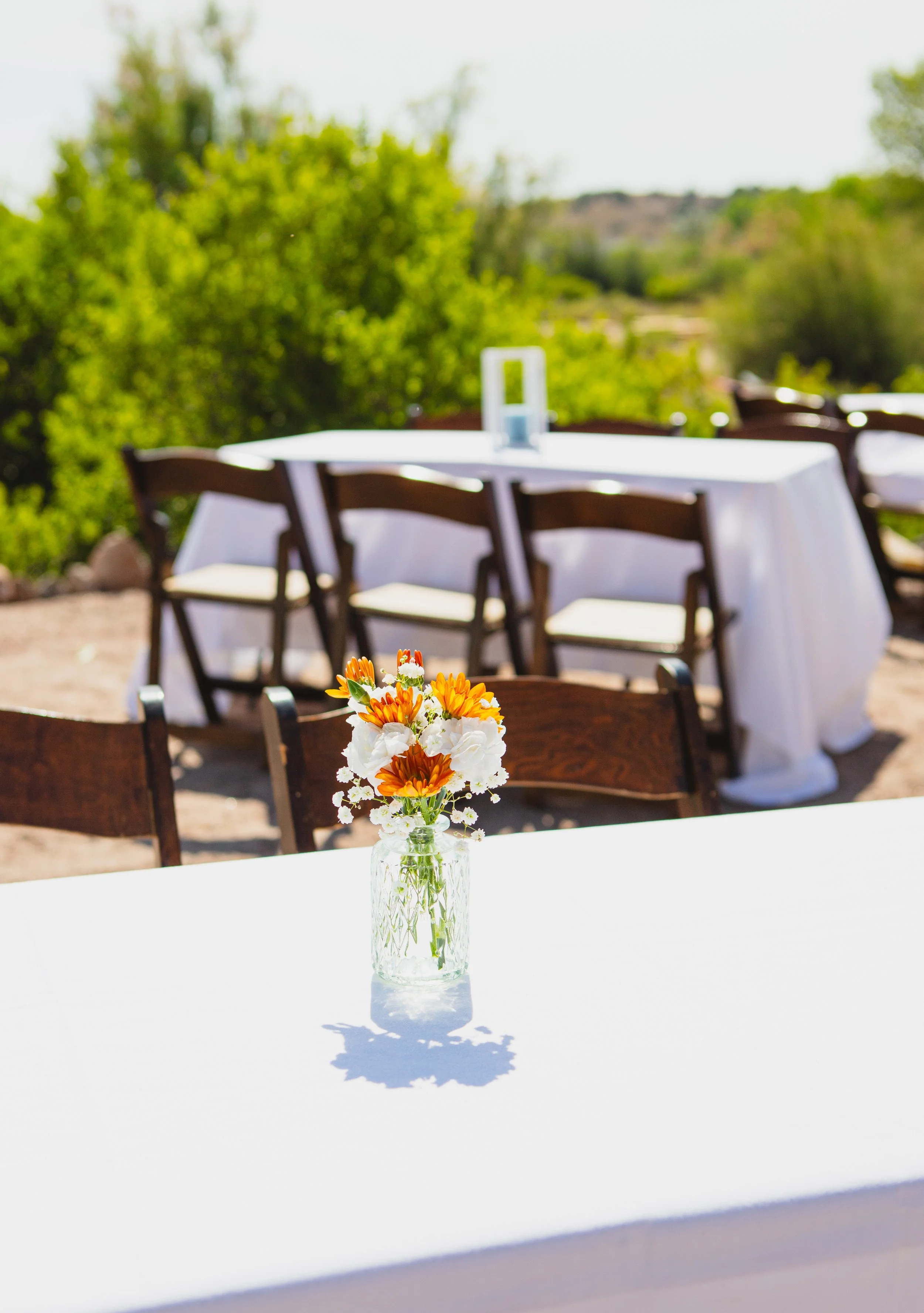 A clear glass vase with orange and white flowers on a white tablecloth, outdoor setting with blurred green trees and brown chairs in the background.