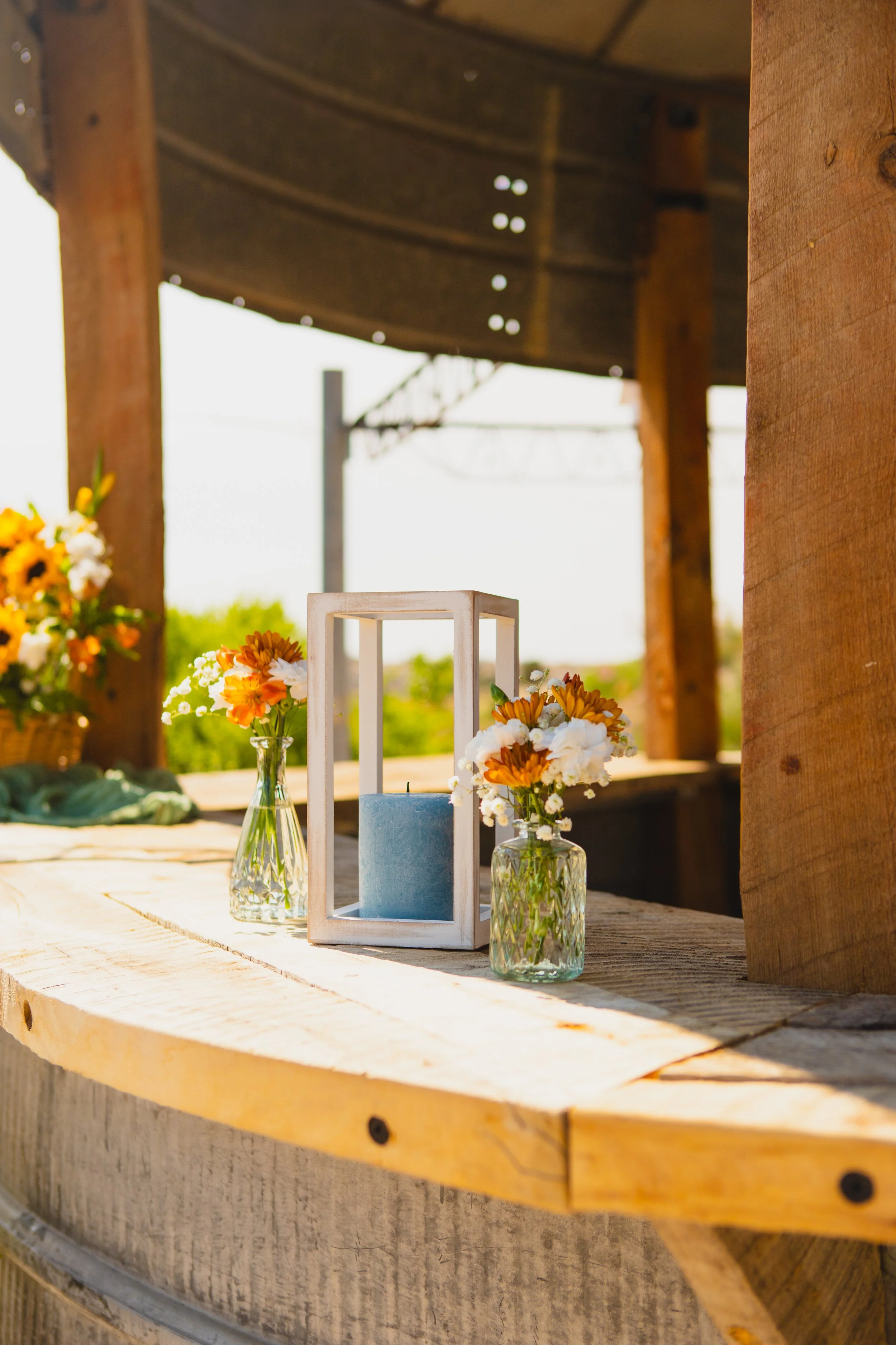 Vases with orange and white flowers, a blue candle inside a white lantern, and a light-colored wooden table on an outdoor deck with a wooden pergola, in a bright sunny setting.