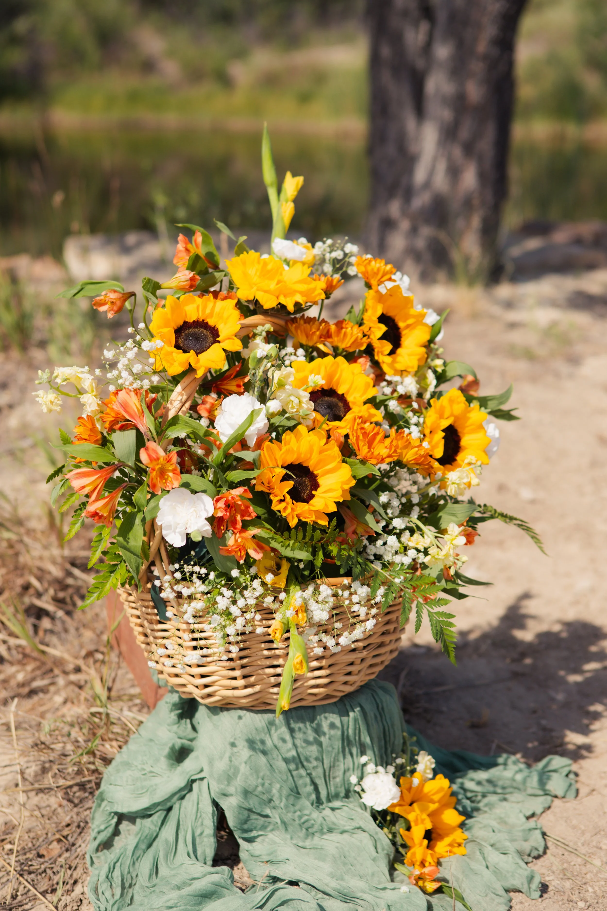 A basket of various bright yellow, orange, and white flowers sitting outdoors on a green cloth with grass and trees in the background.