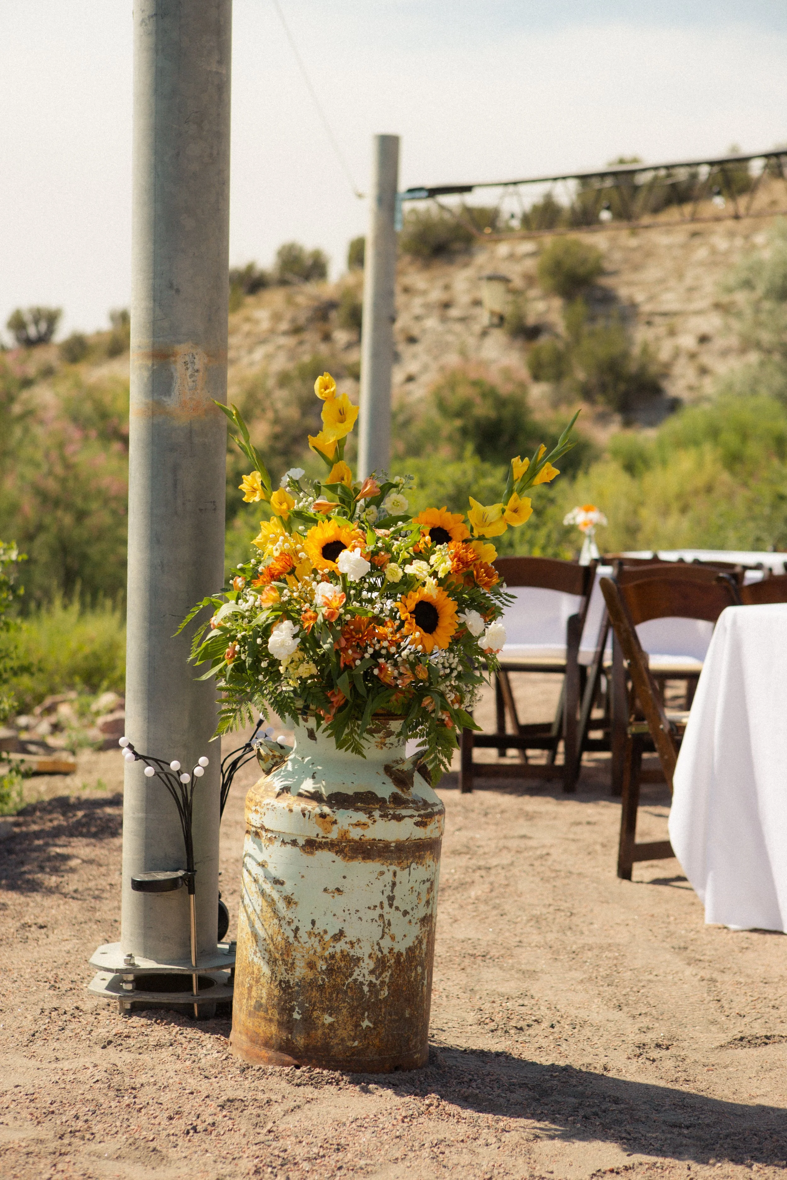 A rustic, weathered ceramic vase filled with a colorful bouquet of yellow sunflowers, orange flowers, and white blooms, set on the ground outdoors next to a tall, rusted metal pipe.