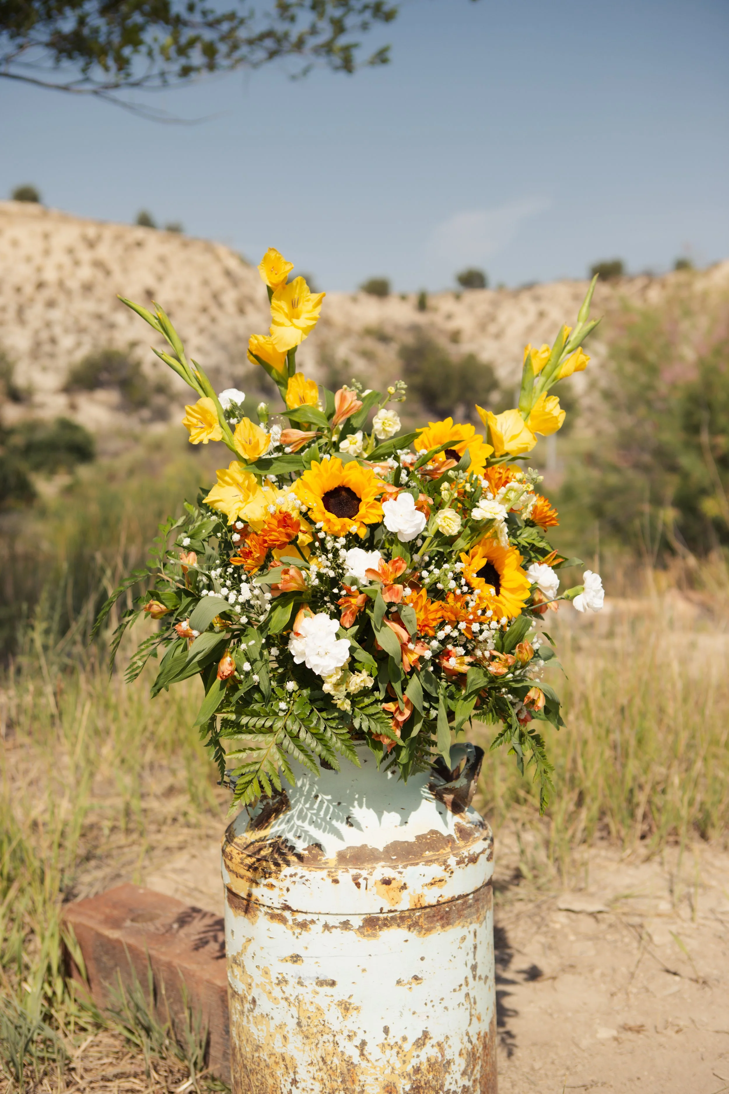 A bouquet of yellow, white, and orange flowers with green leaves in a rustic white vase, placed outdoors on a weathered rusty metal barrel with a background of blue sky, hilly landscape, and sparse trees.