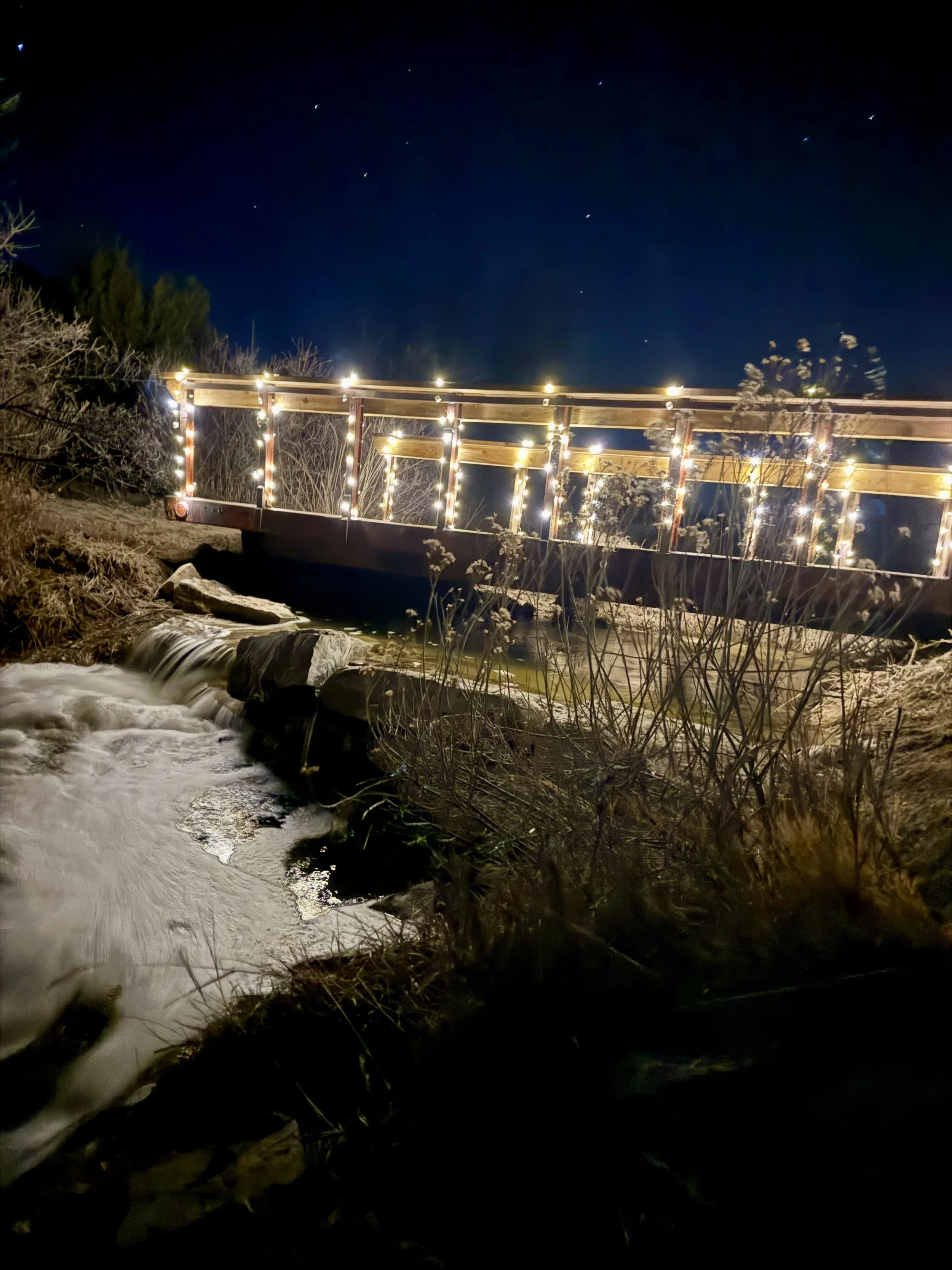 A bridge lit by string lights, the twinkle fairy lights light up the waterfall under the bridge at Lost Pond, a rustic outdoor wedding venue near Pueblo, Colorado.
