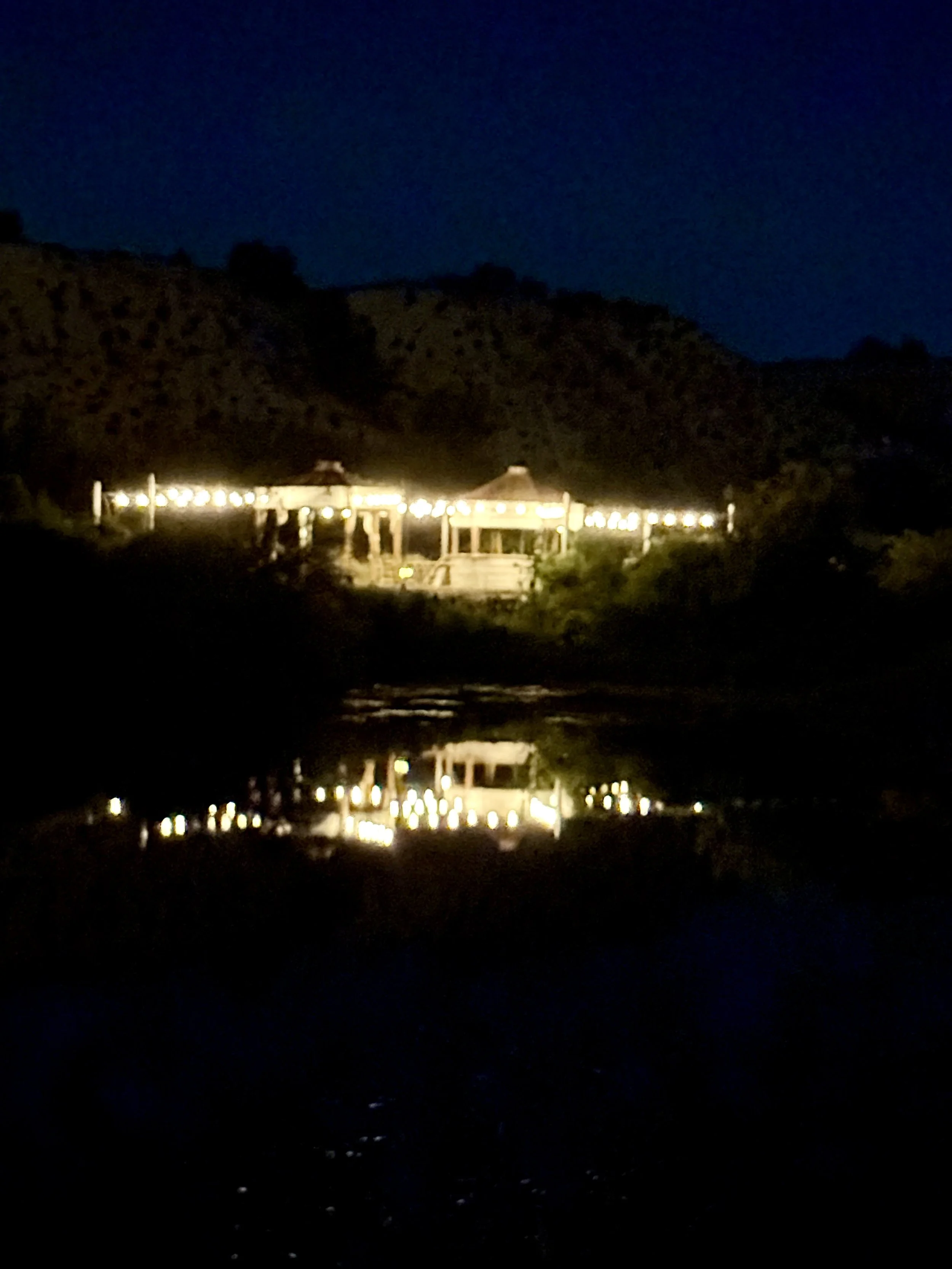 Nighttime scene of a lit-up wedding venue pavilion with string lights, surrounded by darkness and mountains in the background, with reflections on water in the foreground. At Lost Pond outdoor wedding venue near Colorado Springs, Colorado.