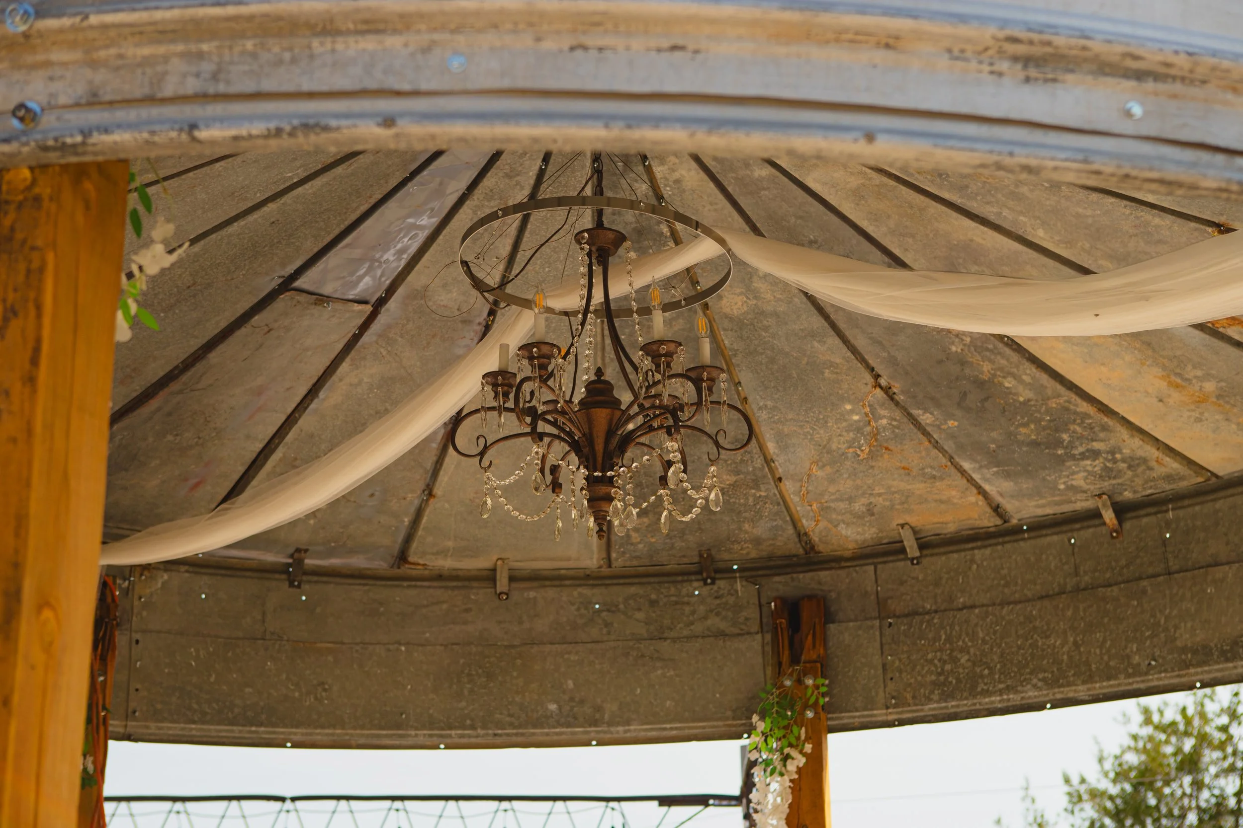 An ornate chandelier hanging from the ceiling of a covered outdoor area with wooden beams and a shingled roof.