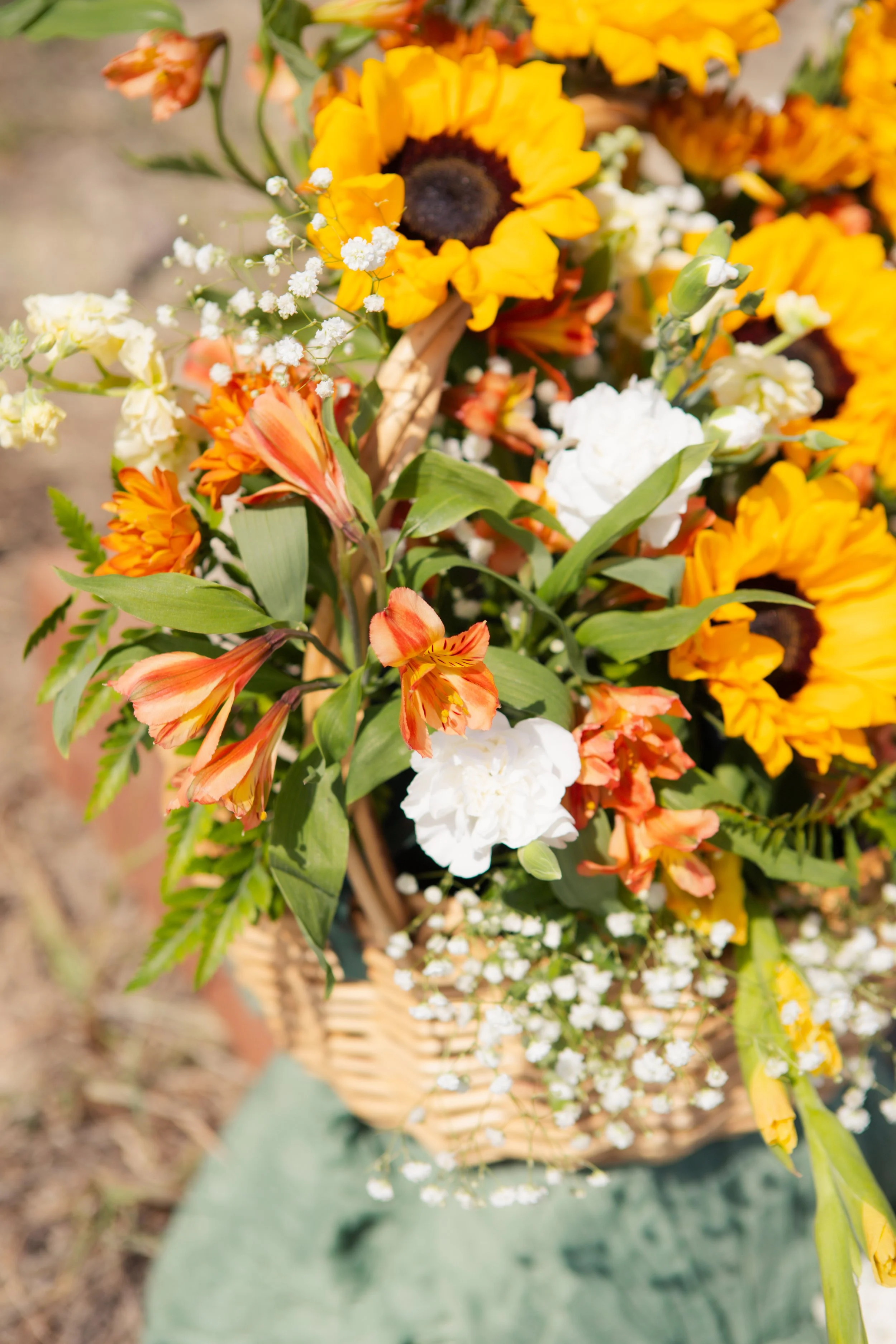 Close-up of a basket filled with colorful flowers, including sunflowers, white carnations, orange lilies, and baby's breath, arranged outdoors.