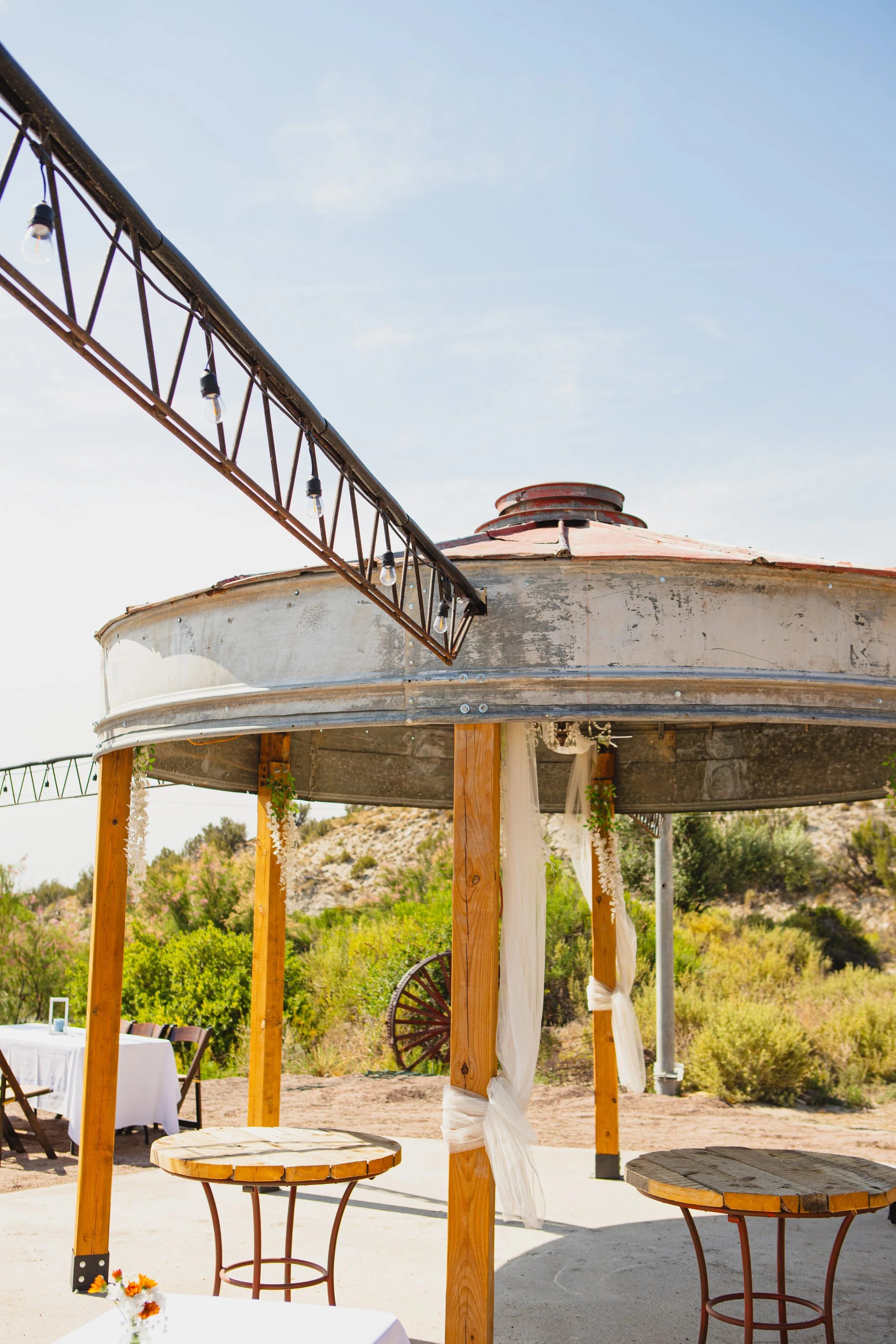 Outdoor dining area with wooden tables, chairs, and an overhead pergola with string lights. Background shows desert landscape and greenery.