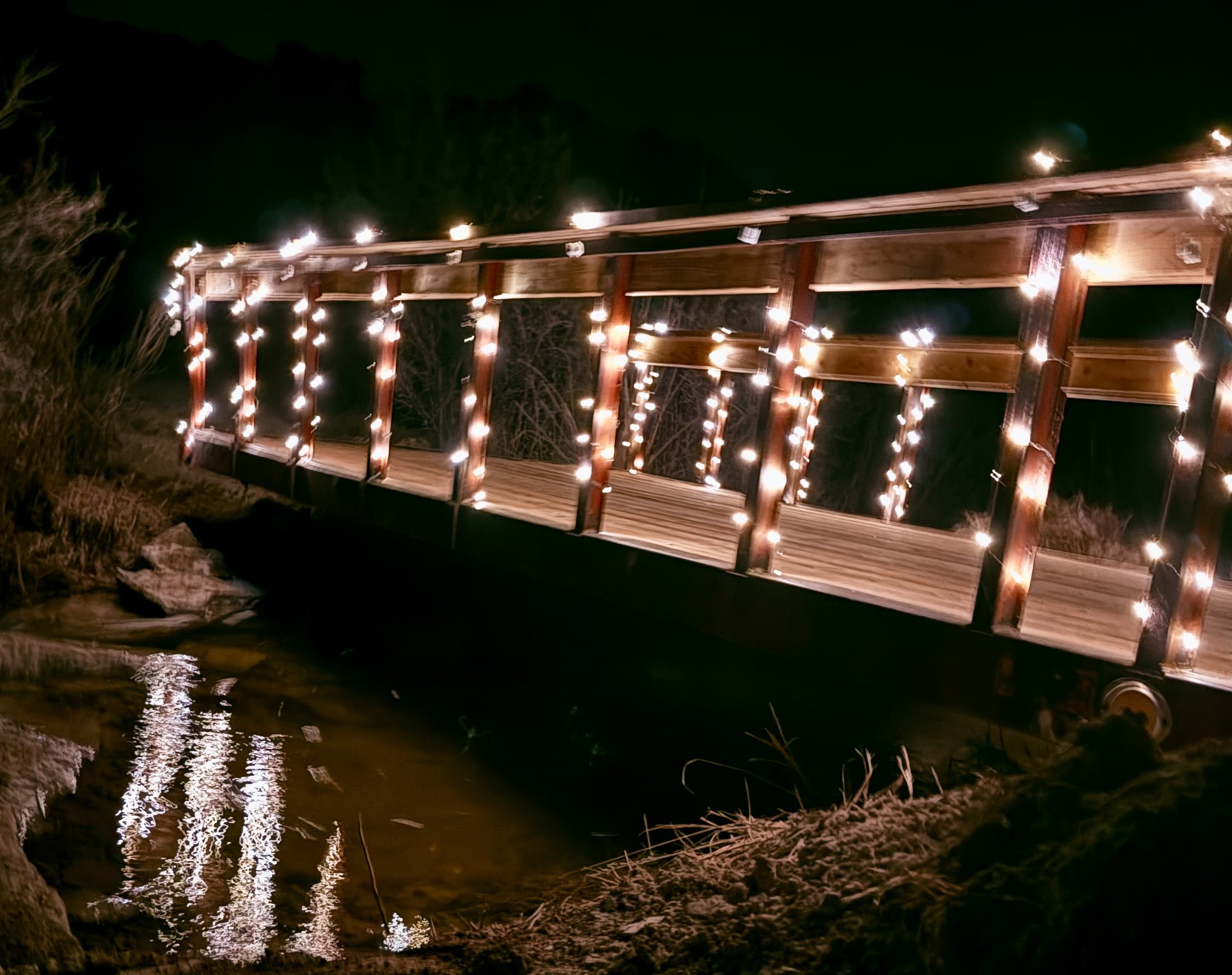 A rustic wooden bridge over a waterfall decorated with whimsical string lights at night, reflected in the water like fairy lights at an outdoor wedding venue near Canon City, Colorado.