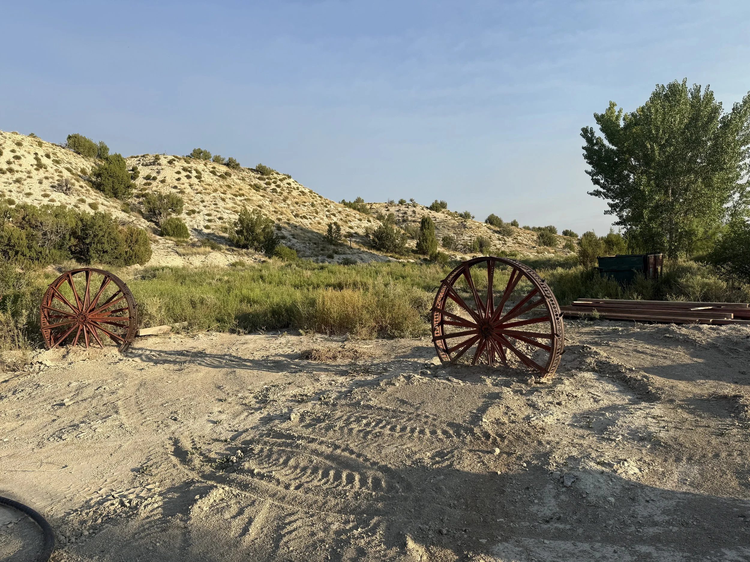 Two old, rusty wagon wheels on dry, sandy ground with green hills and bushes in the background under a blue sky.