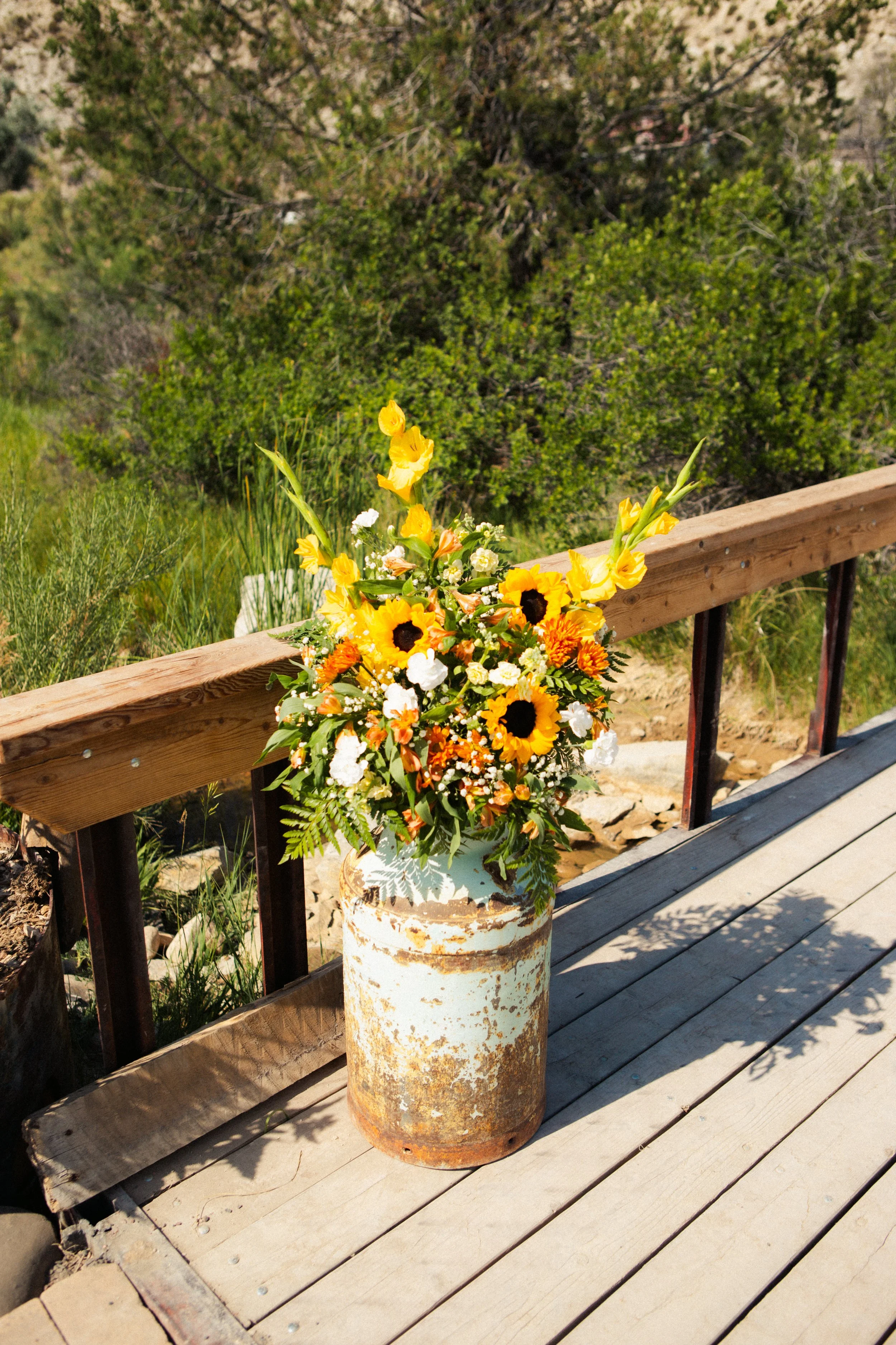 A rustic yellow and white flower arrangement with sunflowers and daisies in a vintage ceramic vase, placed on a rusty metal barrel at a wooden deck with a view of green bushes and trees.
