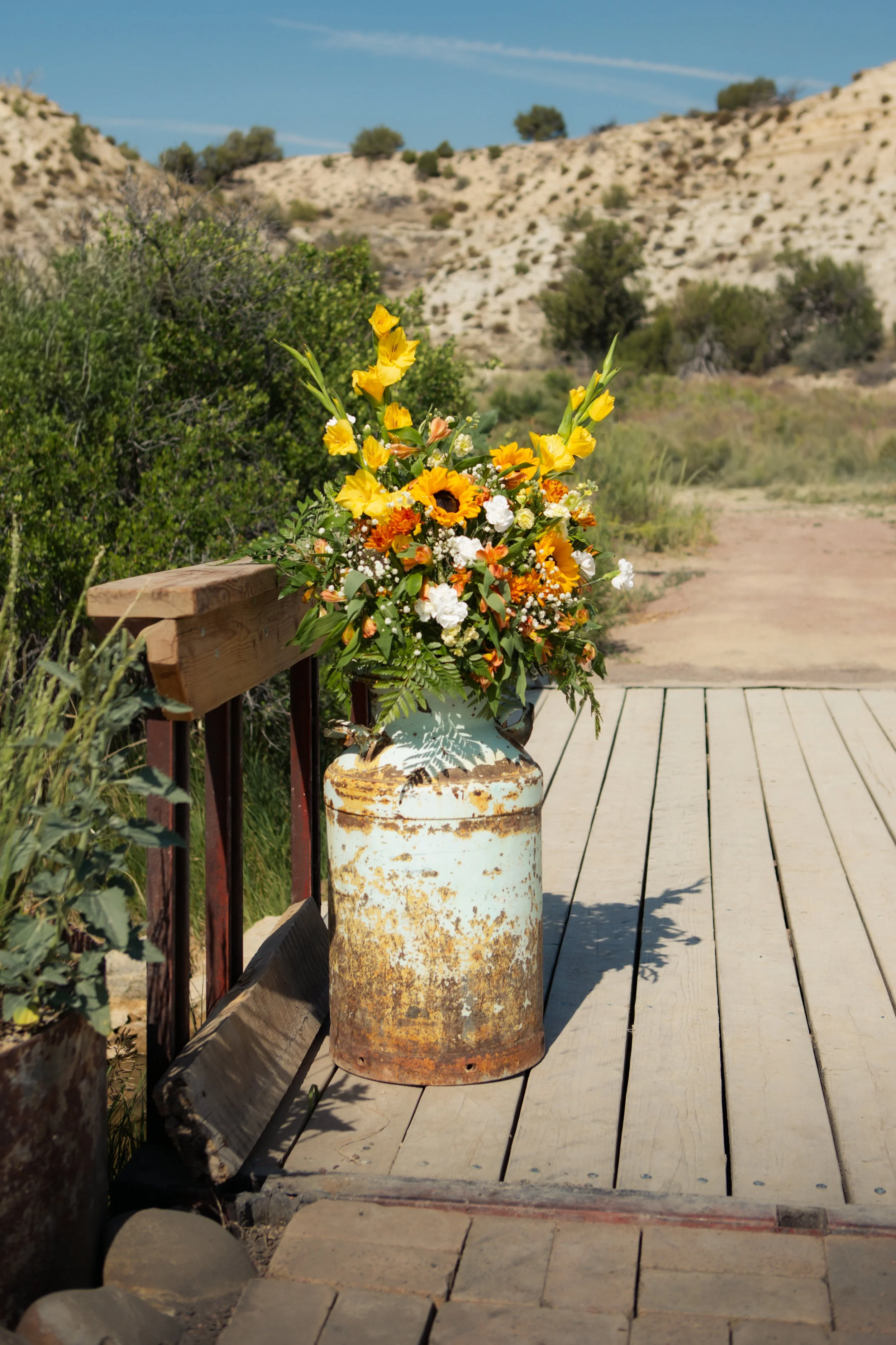 A rustic outdoor scene featuring a large bouquet of yellow, white, and orange flowers in a weathered, rusty metal container, placed on a wooden deck with a scenic desert landscape and hills in the background.
