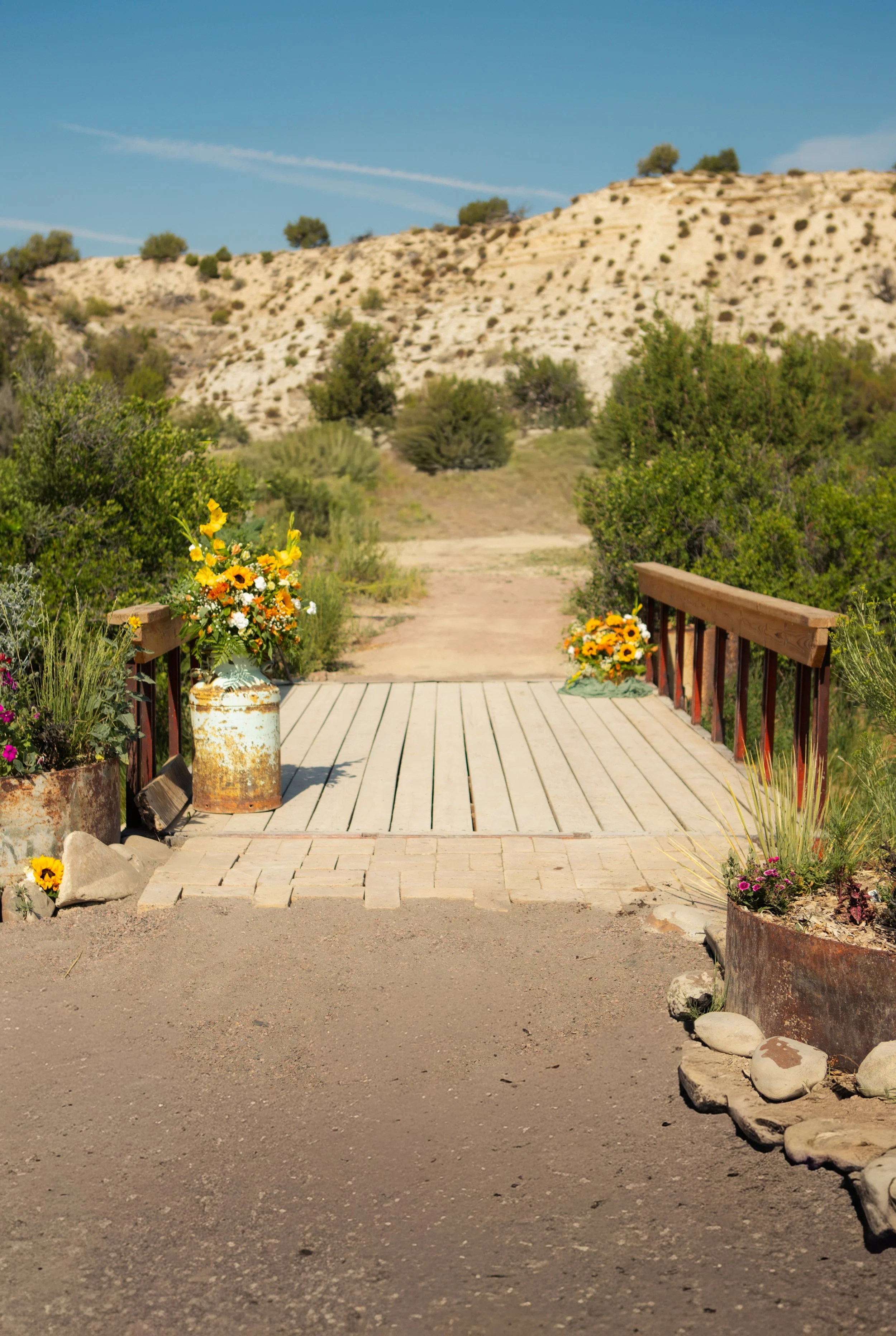 A small wooden bridge decorated with flower arrangements on each side, leading to a dirt path surrounded by greenery and shrubbery, with a hilly, semi-arid landscape and blue sky in the background.