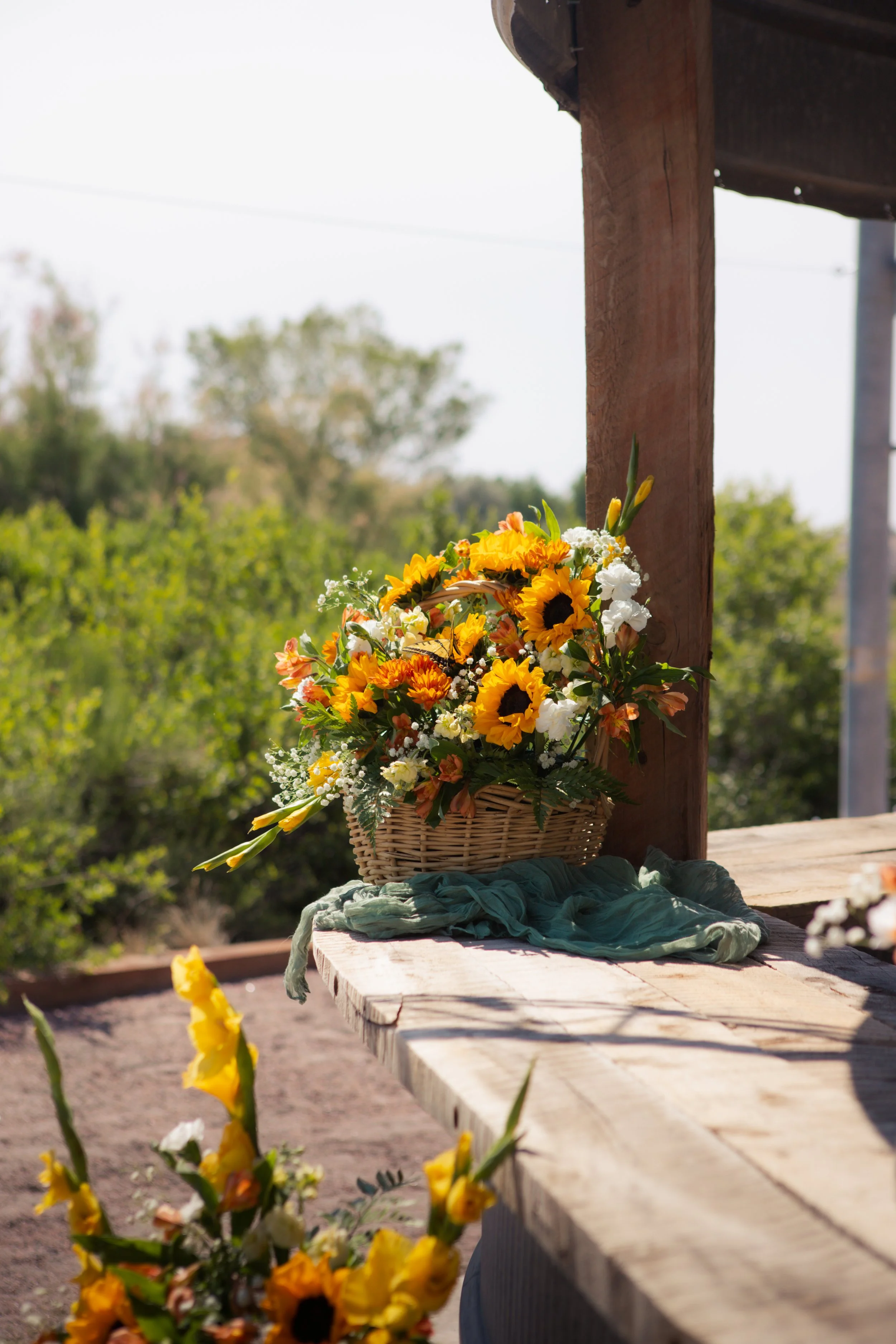 A wicker basket filled with yellow sunflowers and white flowers, placed on a wooden table outside with greenery in the background.