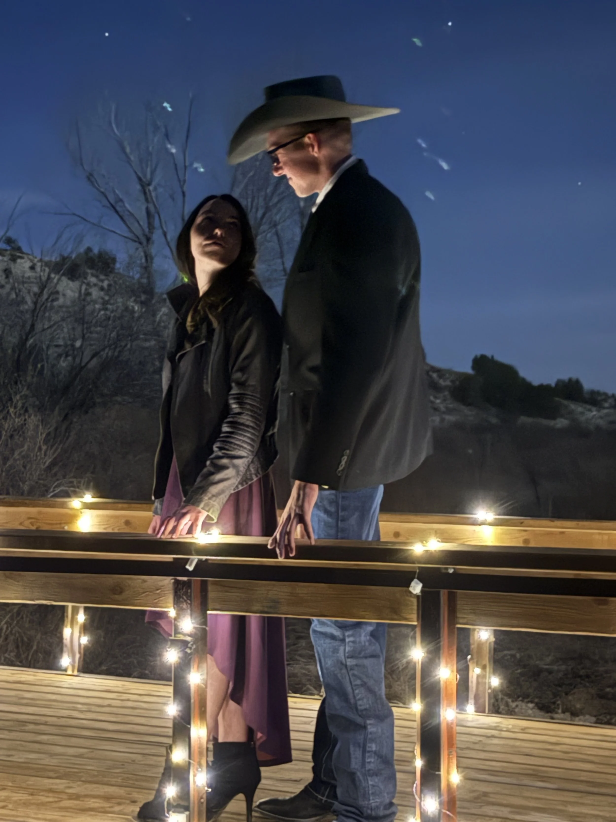 Engaged bride and groom at night on bridge over a waterfall decorated with twinkle fairy lights at night with the starry sky in the background at Lost Pond wedding venue near Pueblo, Colorado.