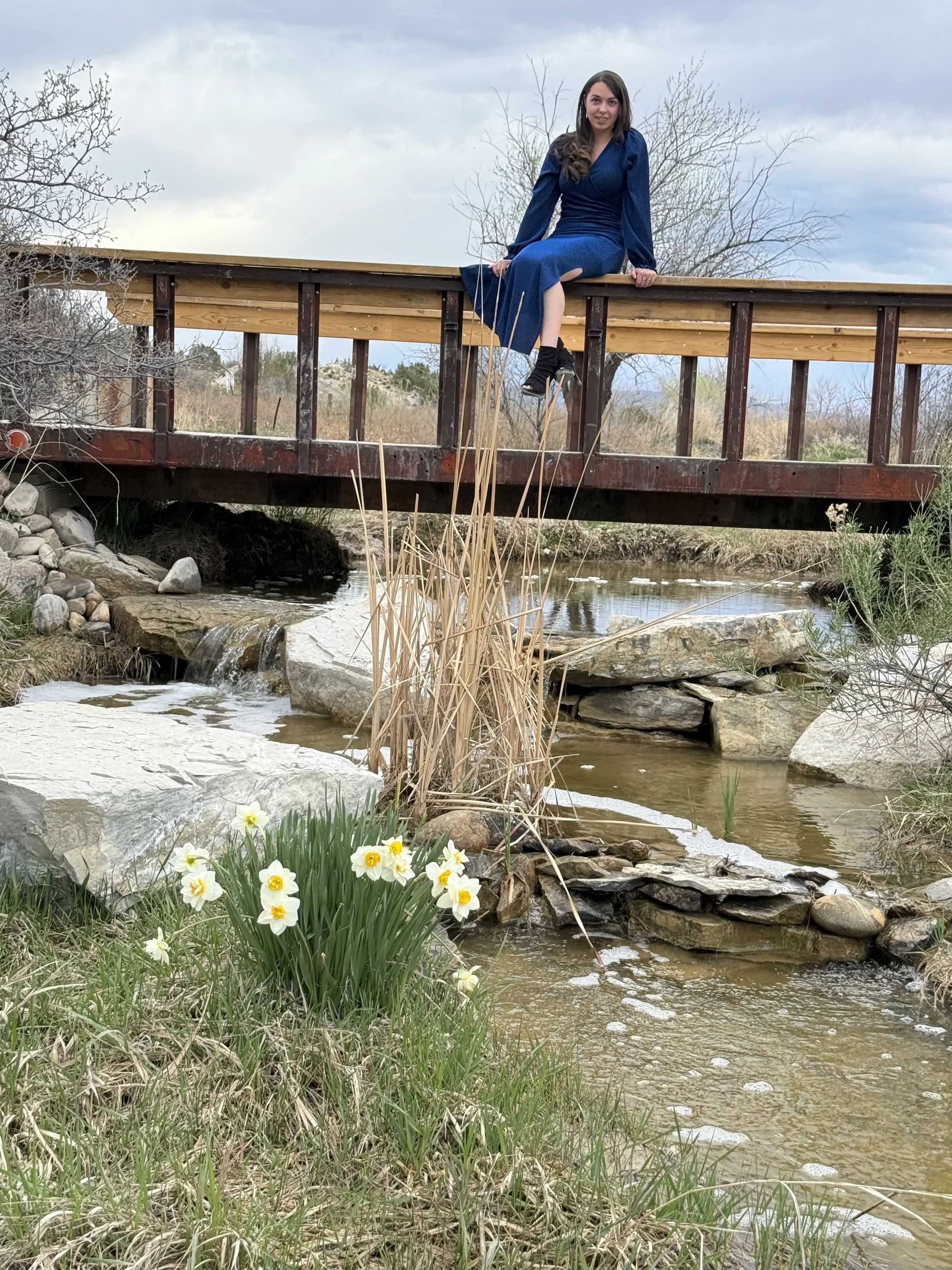 A woman in a blue dress sits on a wooden bridge over a small stream, with blooming yellow and white flowers by the waterfall at Lost Pond wedding venue an event venue with natural water features and wildflowers near Pueblo, Colorado