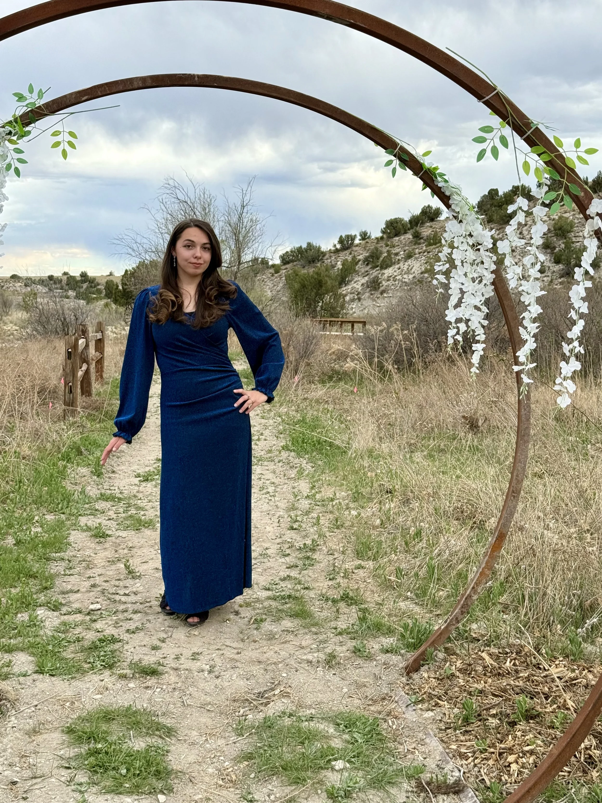 Bride standing on wildflower trail under the moongates with the bridge over the stream in the background. The path through the woods has vines and flowers and grapes that cover the arbor making a whimsical trail to Lost Pond wedding venue near Pueblo