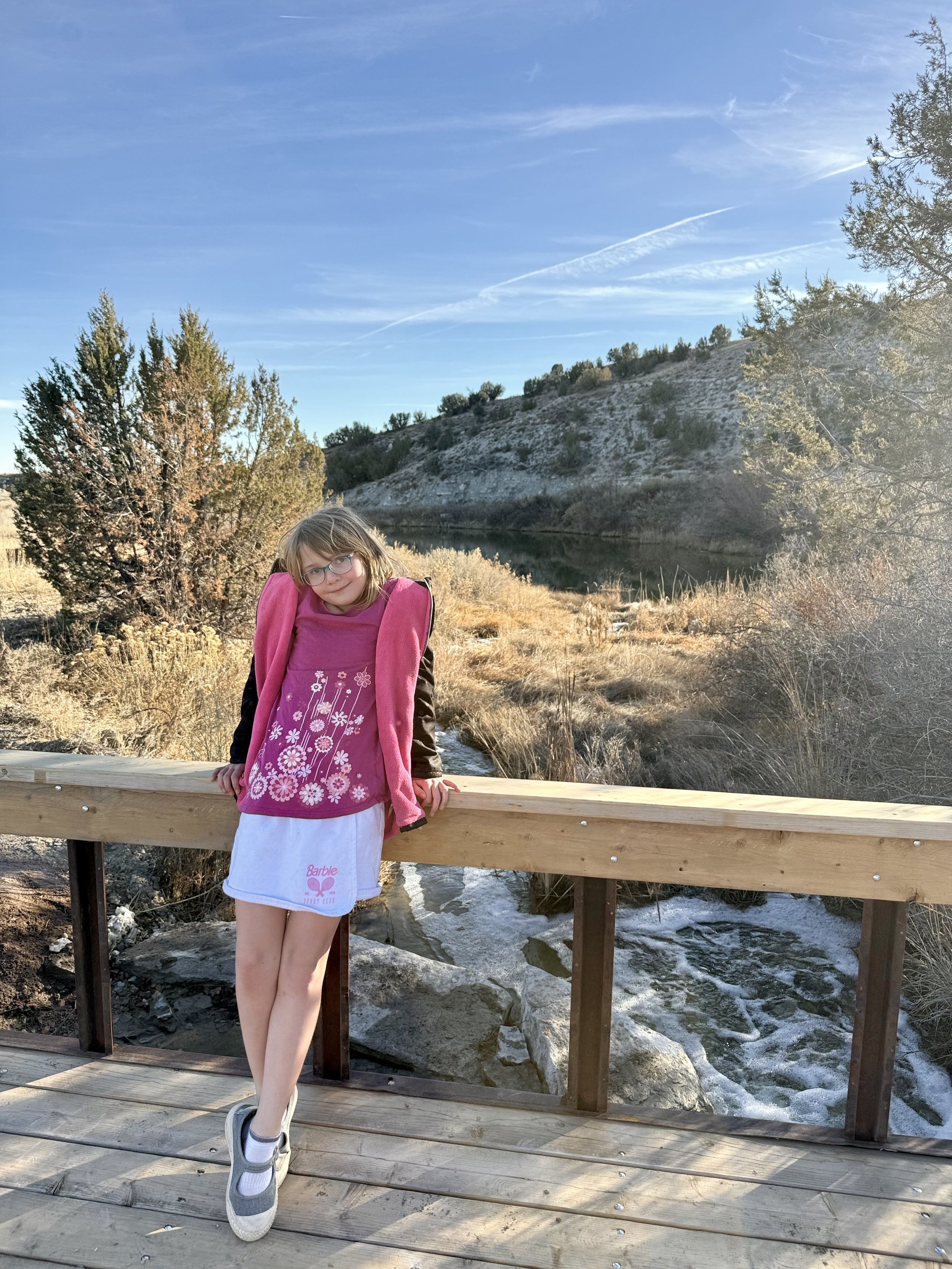 A girl on a rustic wooden  bridge, with a river, waterfall, and mountains in the background at Lost Pond outside wedding venue near Pueblo, Colorado.