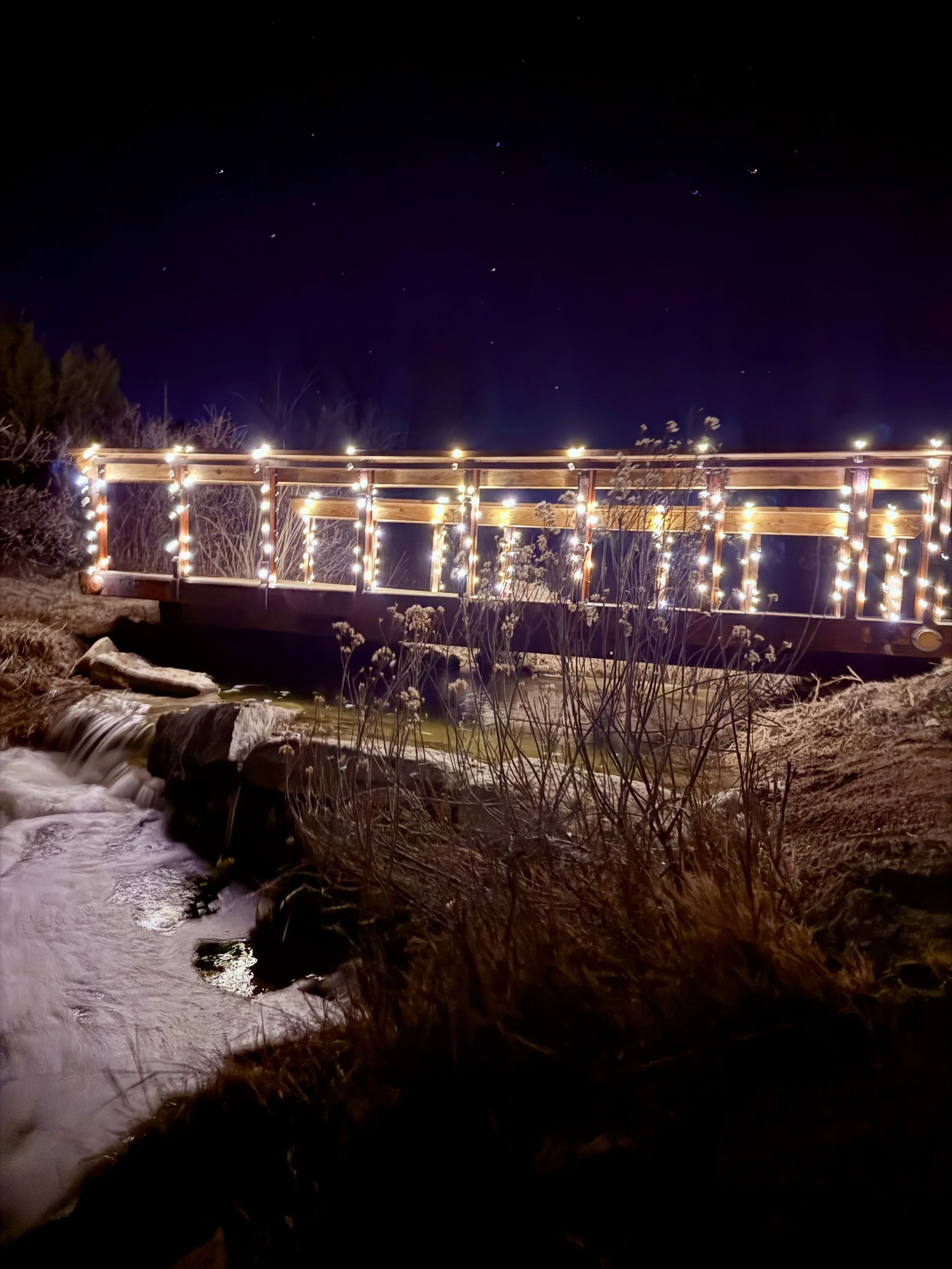 Bridge over the waterfall lit up with twinkle lights at night at Lost Pond wedding venue near Canon City, Colorado.
